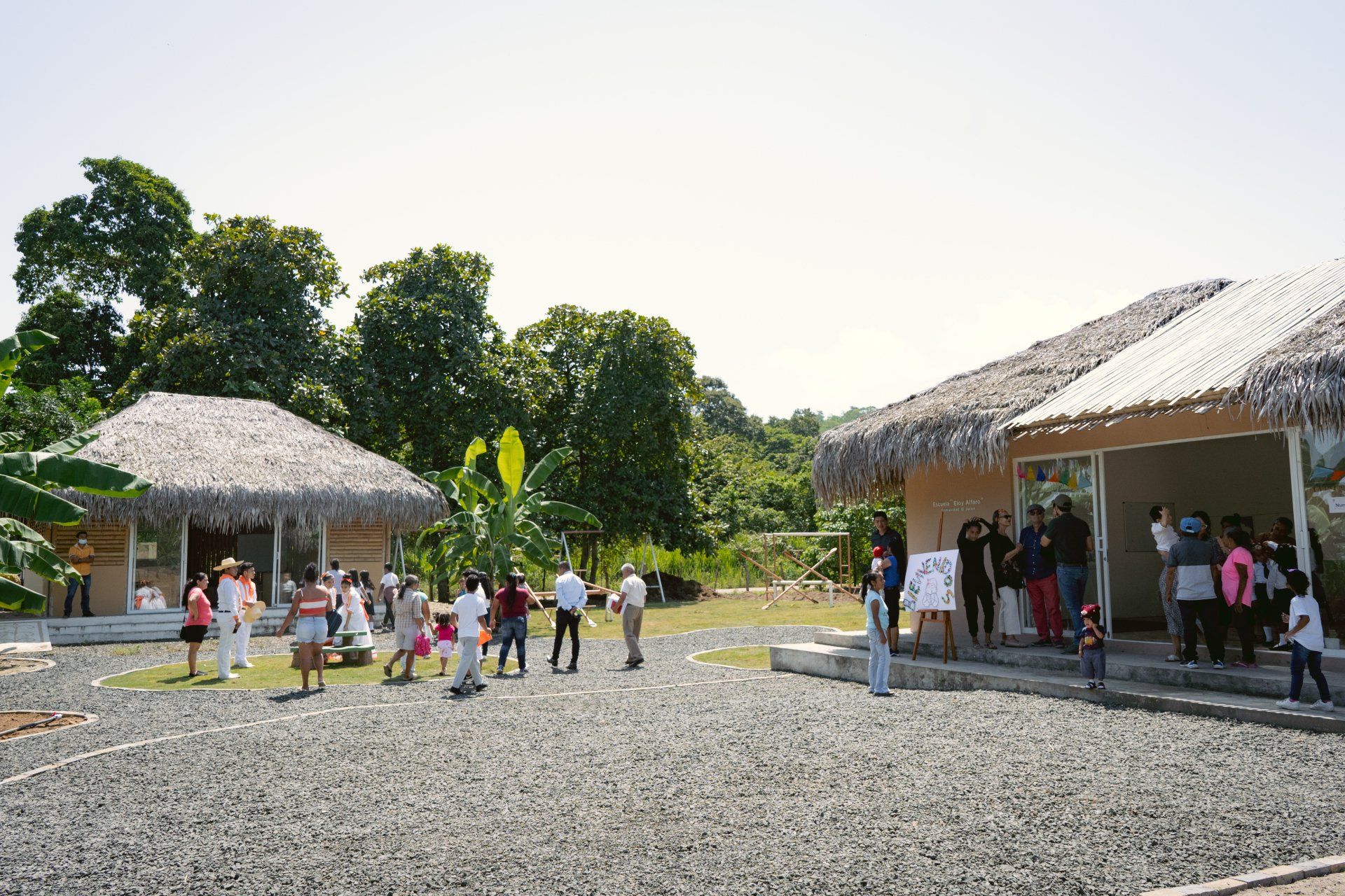 People gather outside two thatched-roof buildings in a sunny outdoor setting.