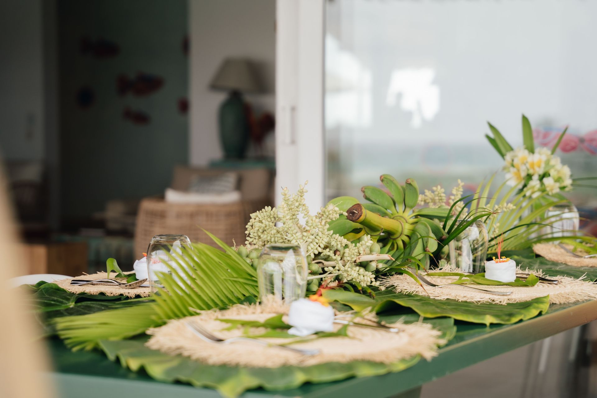 A tropical-themed table setting with green foliage, white flowers, candles, and glass jars.