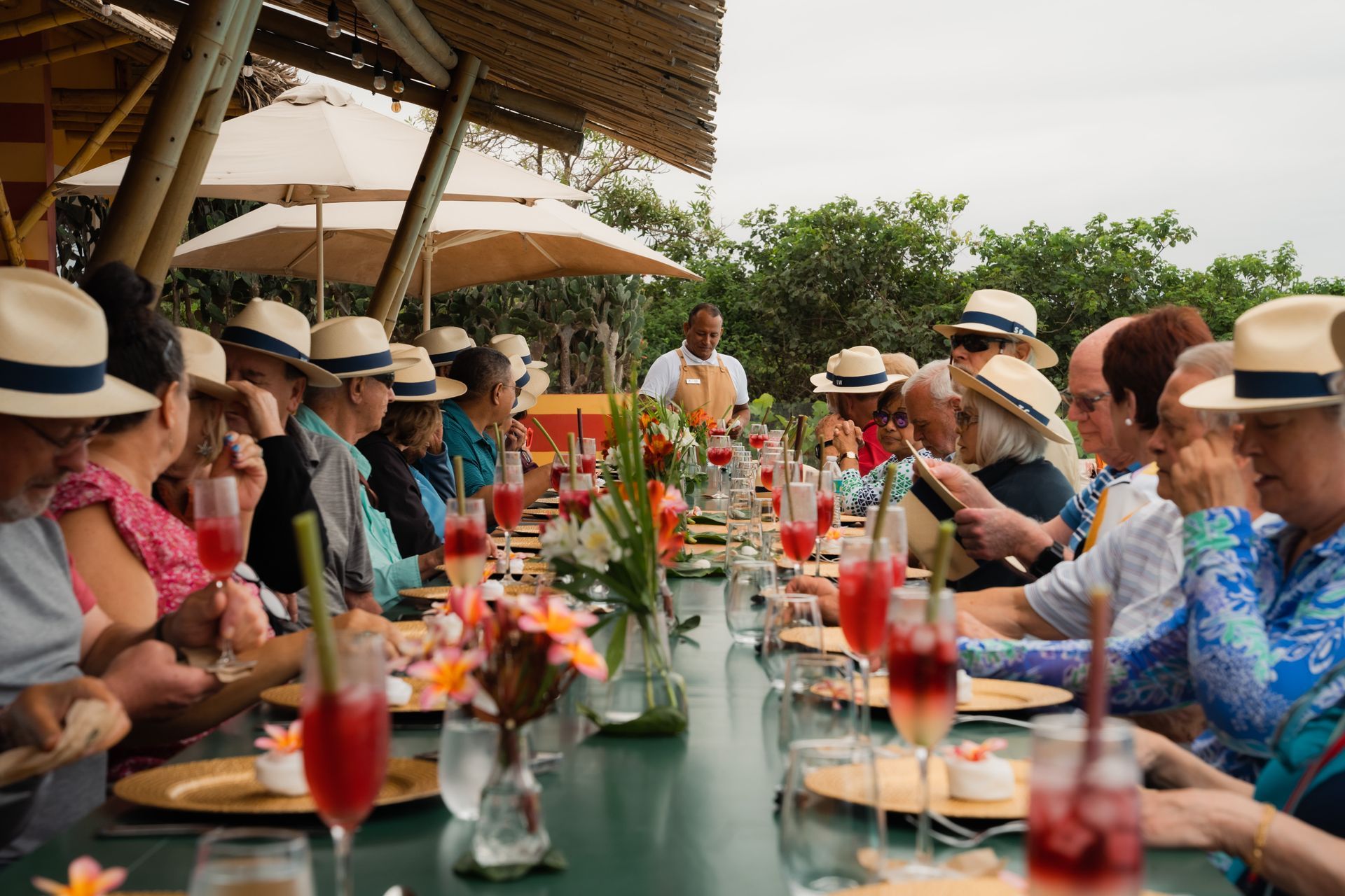 People in hats at a long outdoor table, dining. Table has food, drinks, flowers.