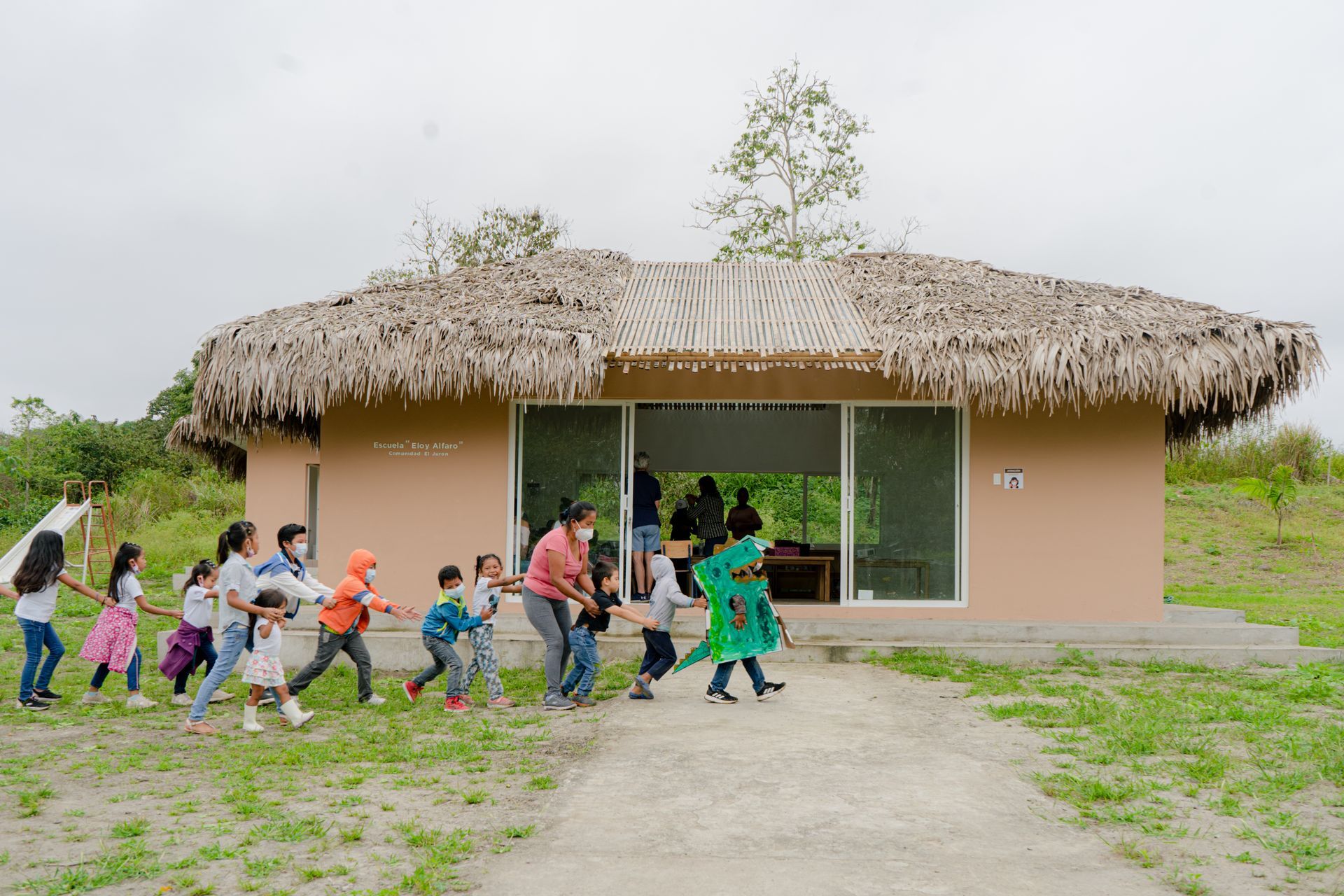 Children and adults walk into a tan building with a thatched roof; green grass surrounds it.