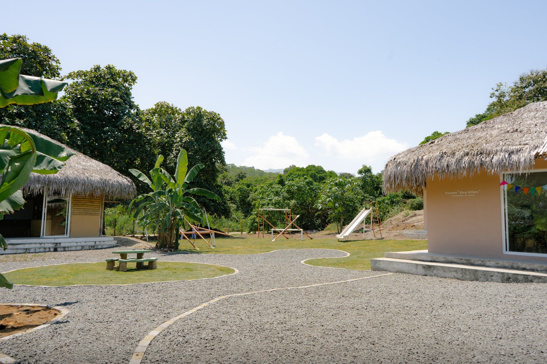 Two huts with thatched roofs, gravel path, and playground in a tropical setting.