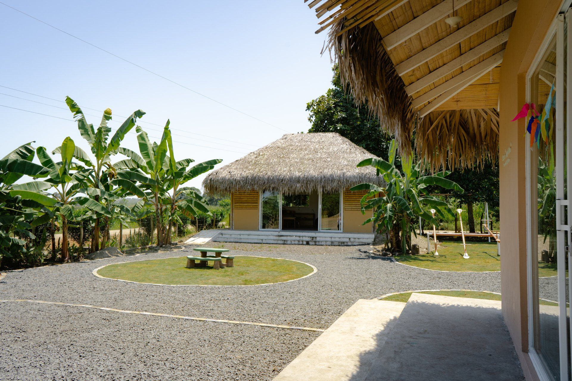 Tropical scene: a round gravel yard with a thatched-roof building, surrounded by banana trees, and a beige building on the right.