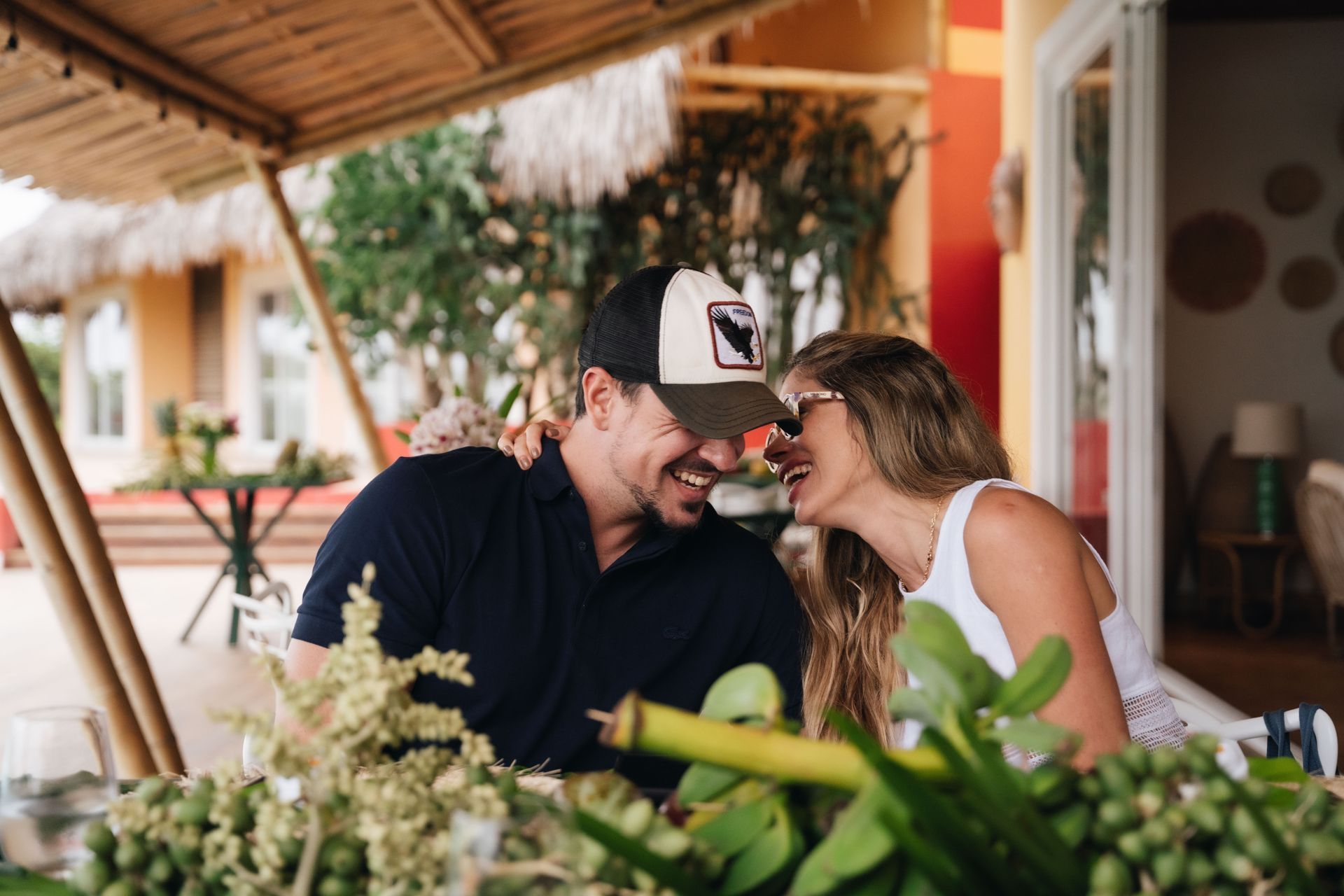 Couple laughing together outdoors, near a table with greenery, under a straw roof.