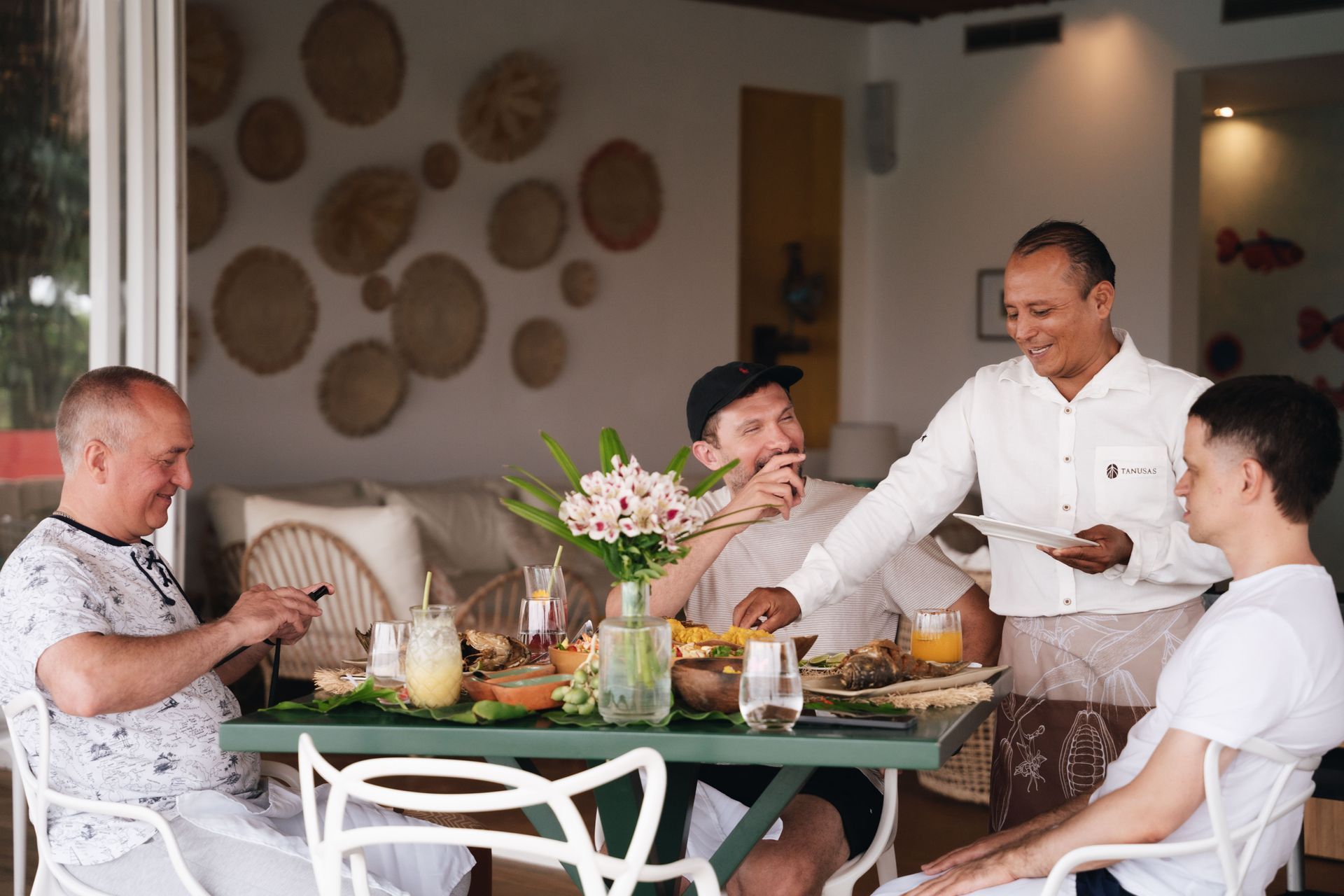 Four people eating at a table; waiter serves. Light-filled room; neutral colors, woven wall decor, floral centerpiece.