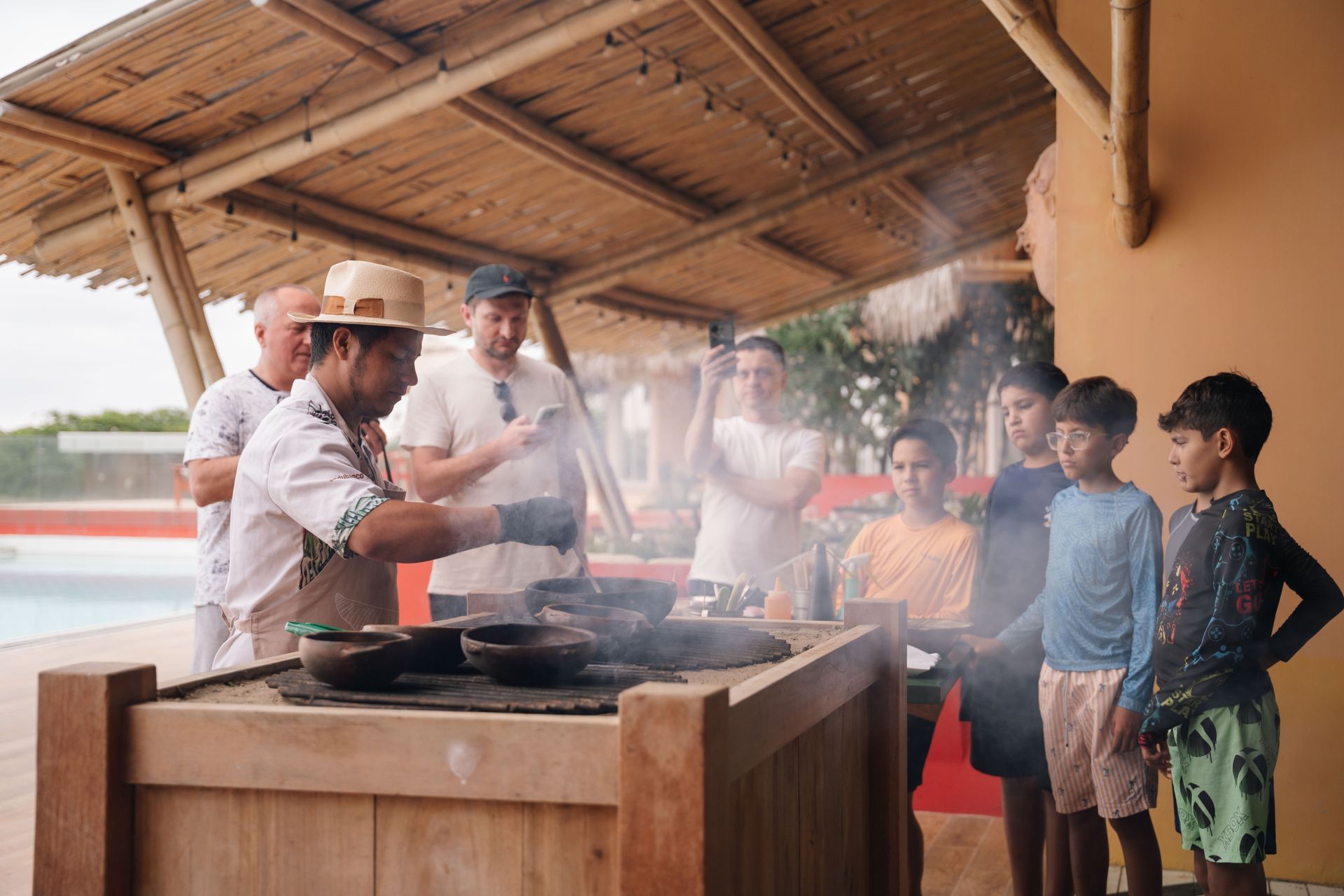 Chef cooking outdoors for a group of people, including children. Smoke rises. Pool visible.