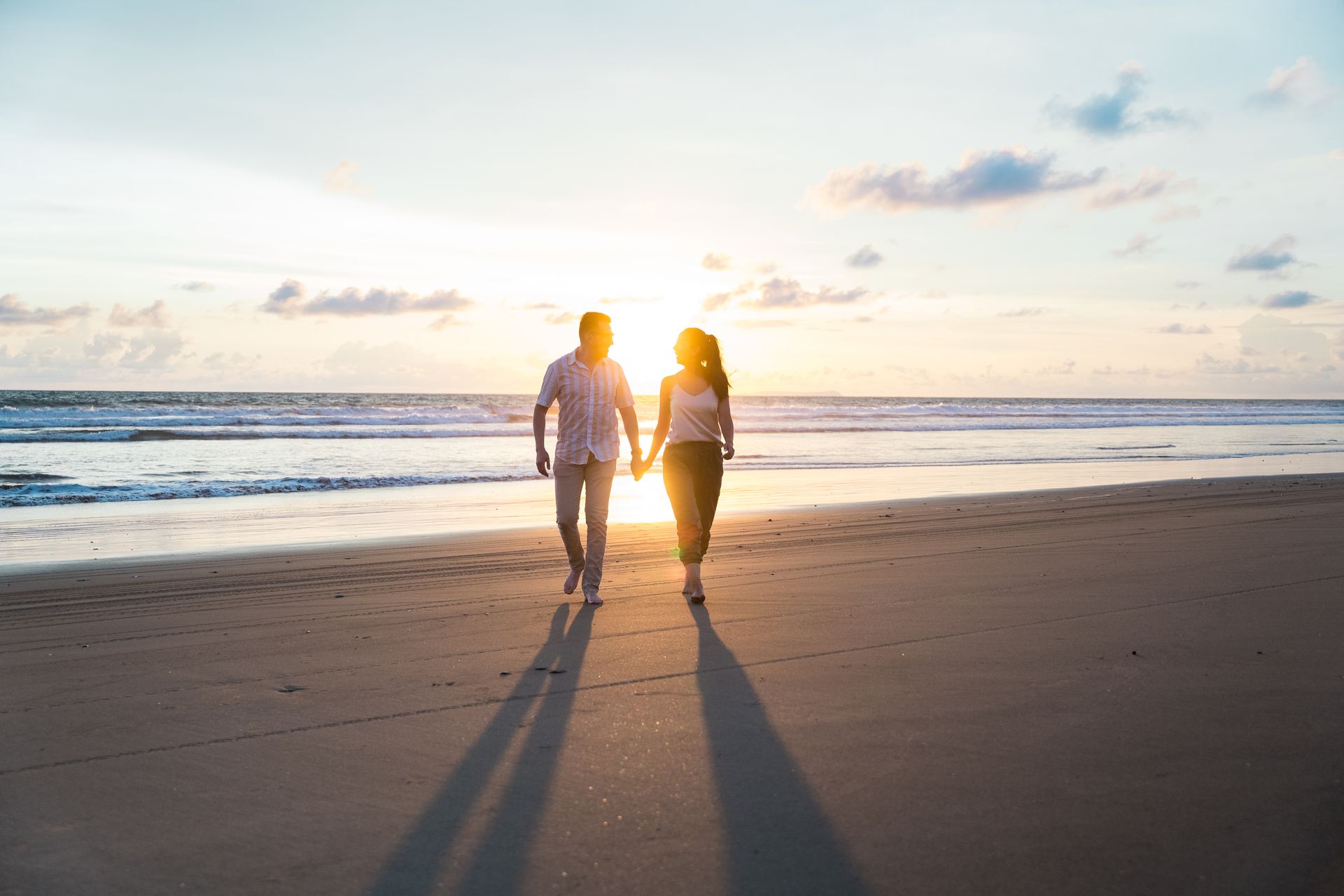 Couple holding hands walking on a beach at sunset, silhouetted by golden light.