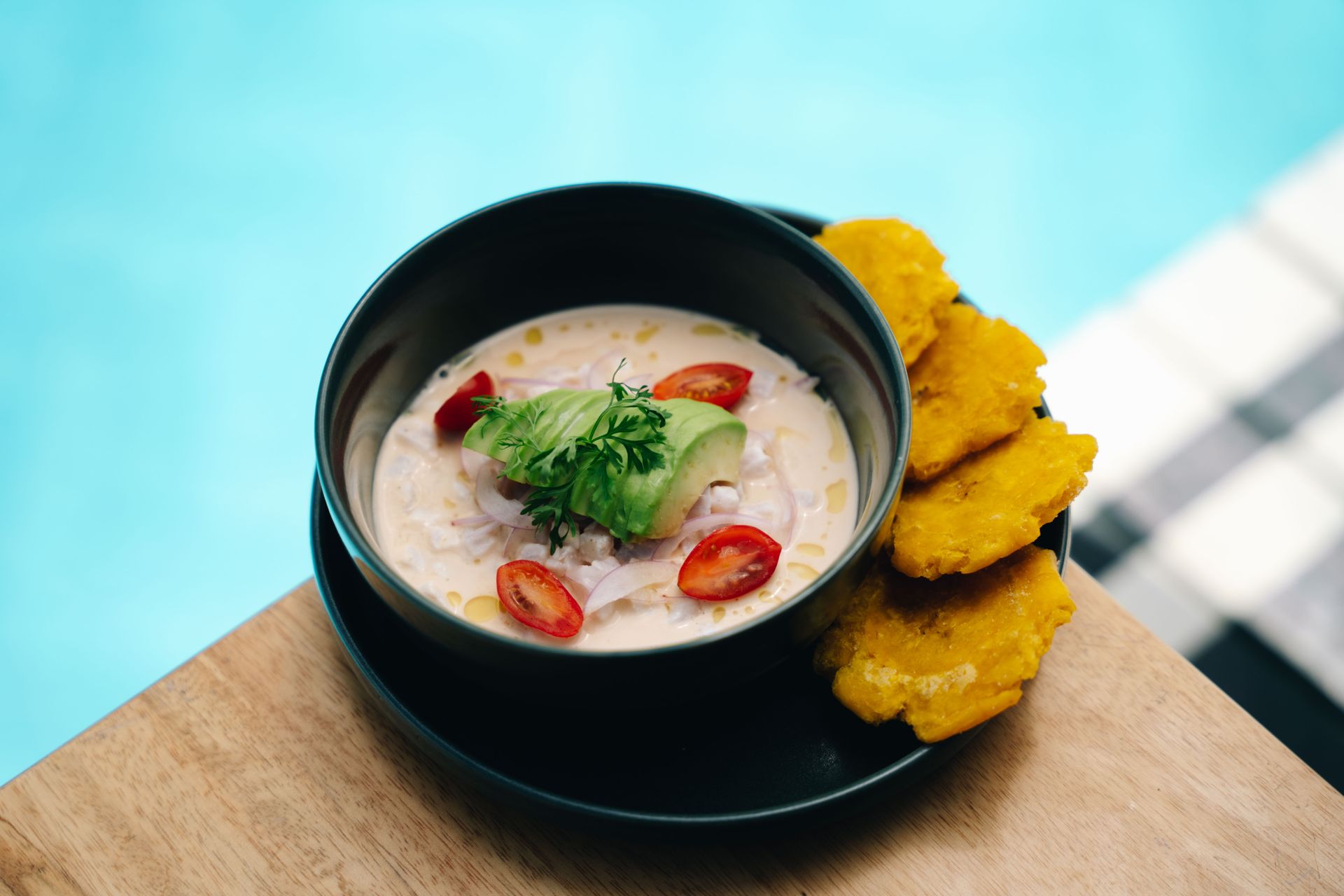 Ceviche with avocado, tomatoes, and plantain chips on a wooden table near a pool.