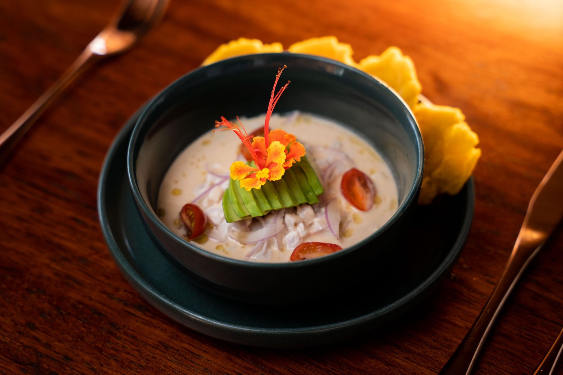 Ceviche in a teal bowl, topped with avocado and flowers, served with plantain chips on a wooden table.
