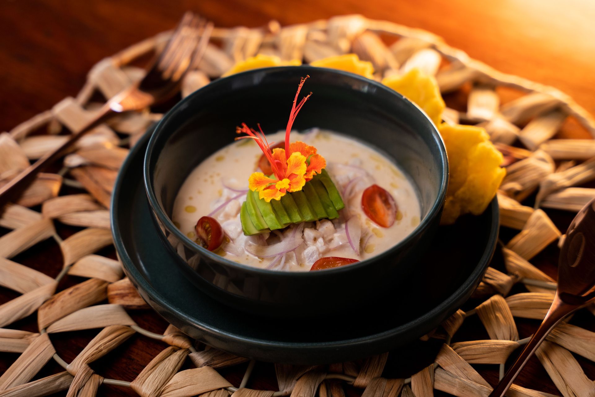 Bowl of creamy soup with avocado, tomatoes, and flowers, served on a woven placemat.