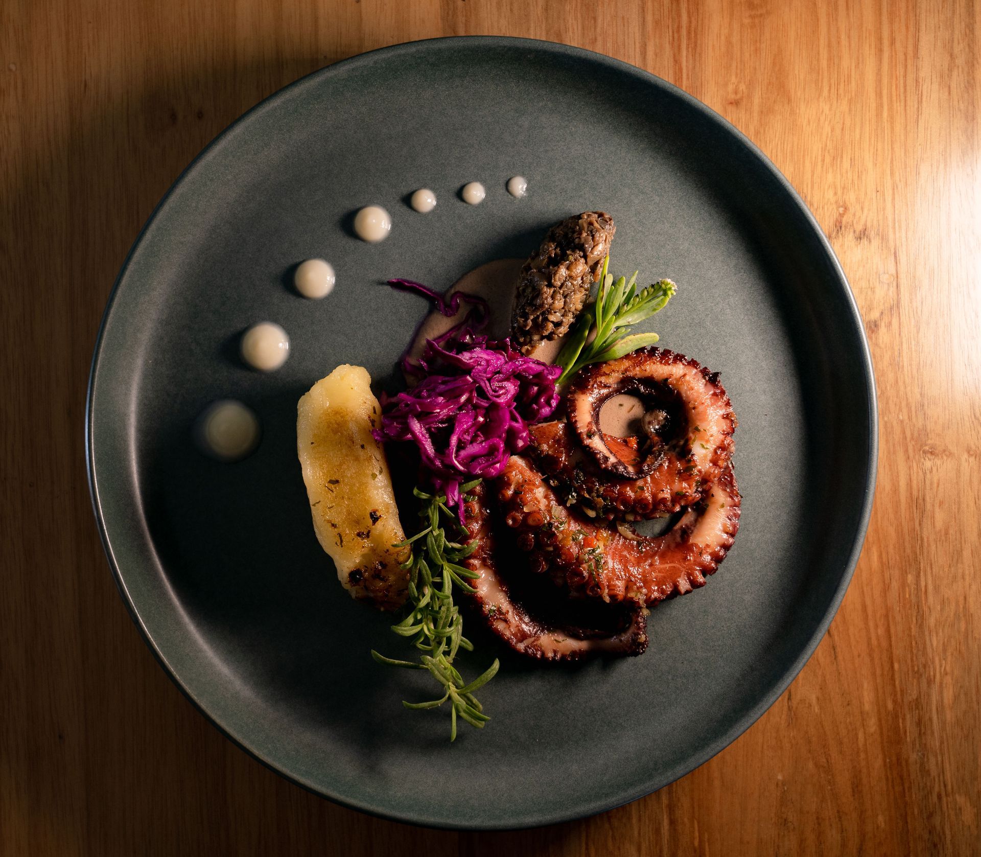 Grilled octopus dish with red cabbage, sauce dots, herbs, on a grey plate, wood table.