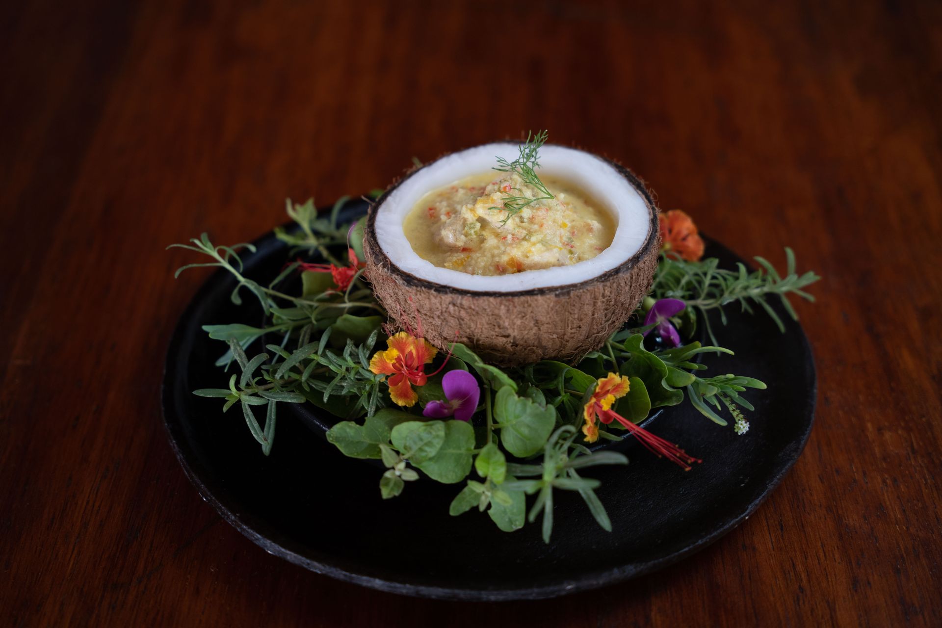Coconut shell filled with yellow food, garnished with flowers and herbs on a black plate.
