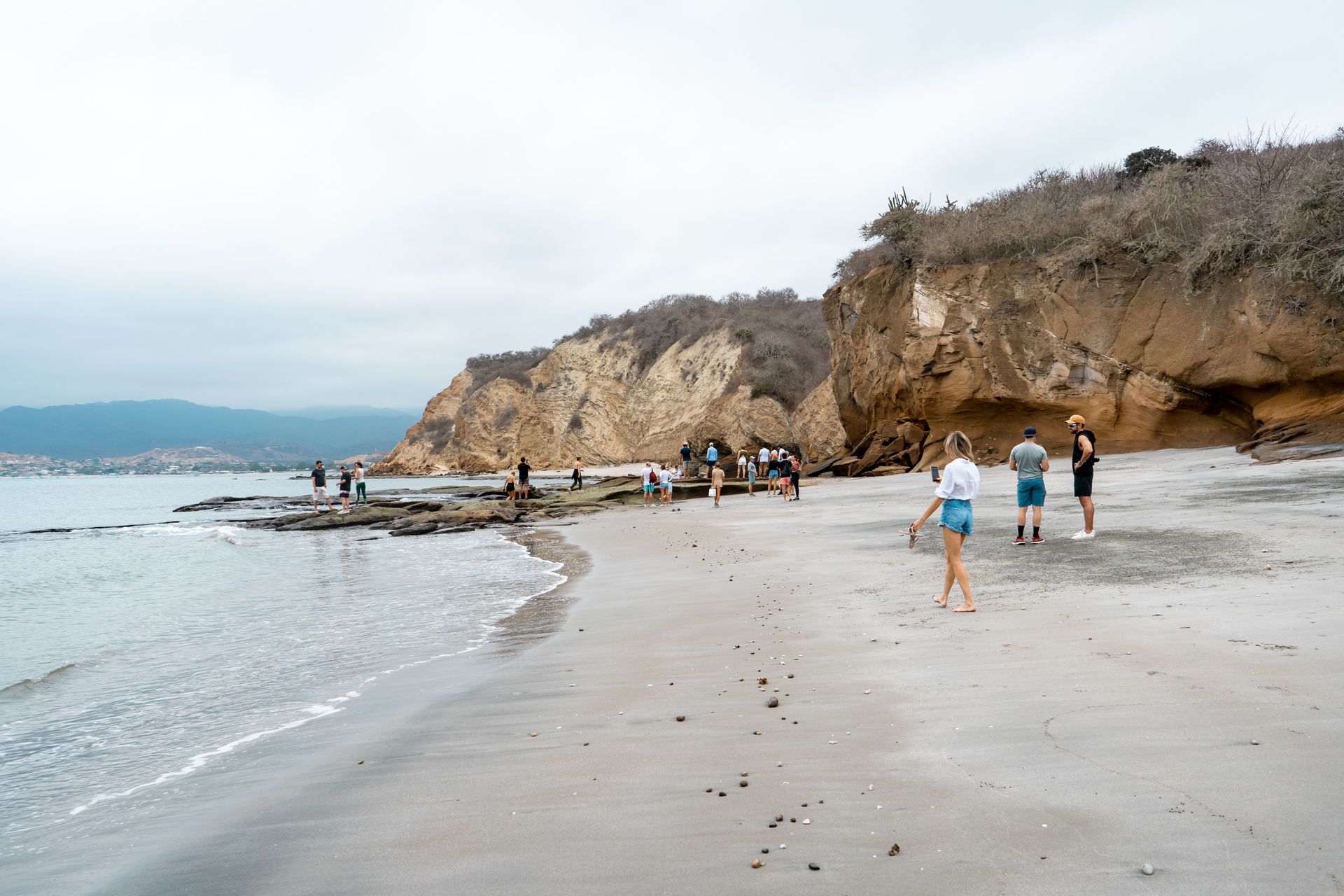 Beach scene with people near rocky cliffs and water under a cloudy sky.