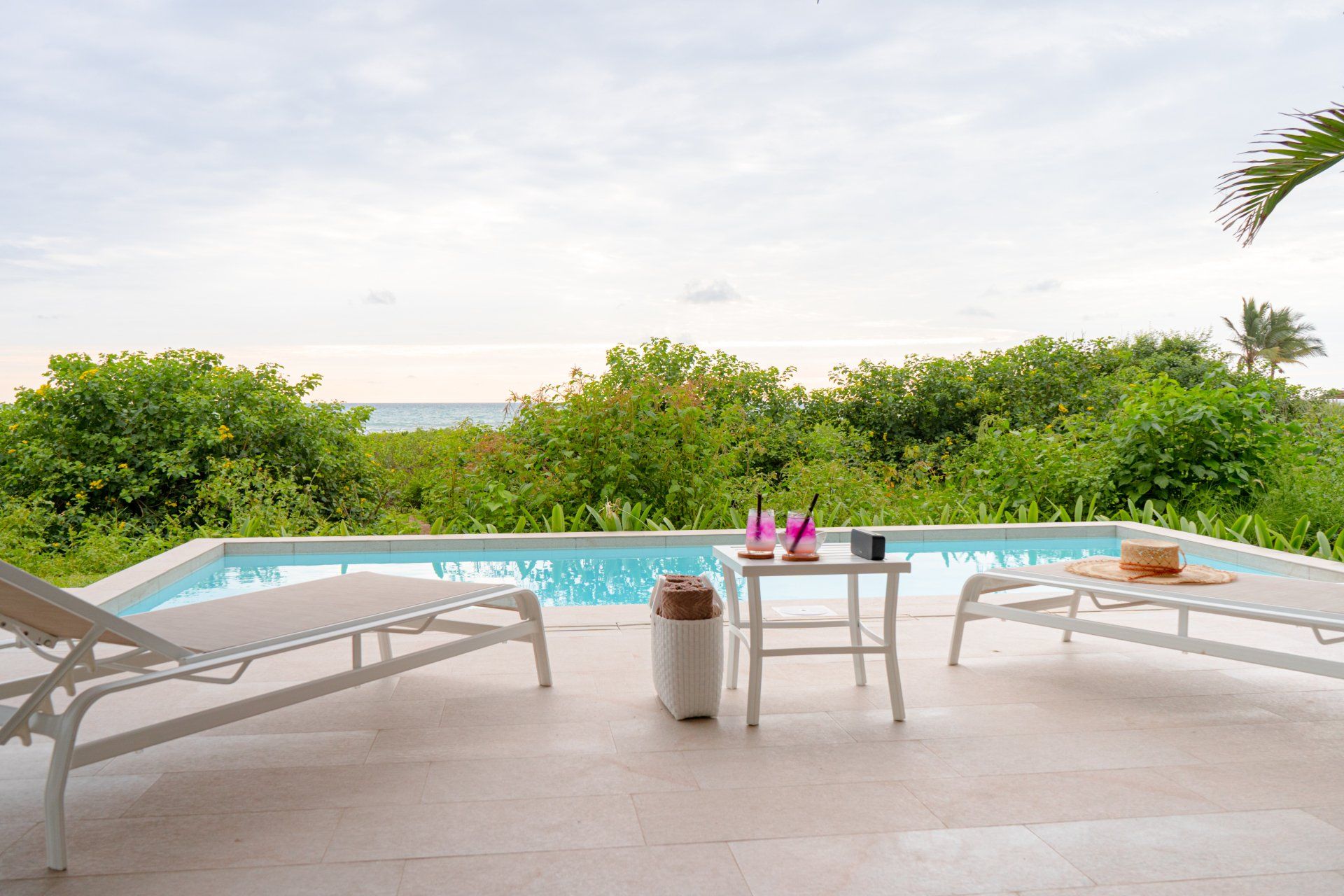 Poolside scene: two lounge chairs, small table with drinks, overlooking lush greenery and ocean under a cloudy sky.