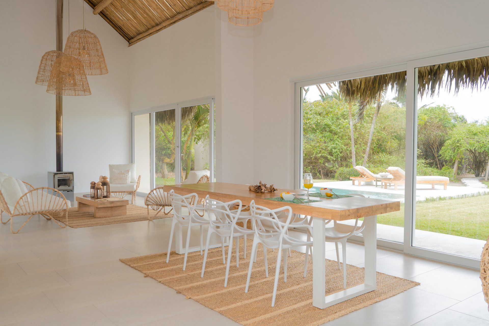 Bright dining area with long wooden table, white chairs, and large windows overlooking greenery.