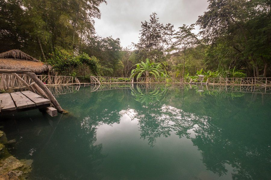 Tranquil pool of turquoise water surrounded by lush greenery, wooden dock, and a thatched roof structure.
