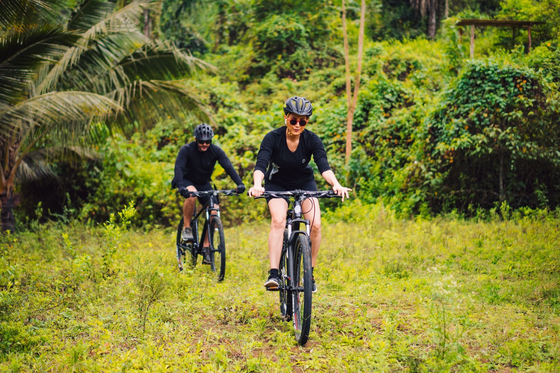Two people bike through a grassy path in a lush green forest.