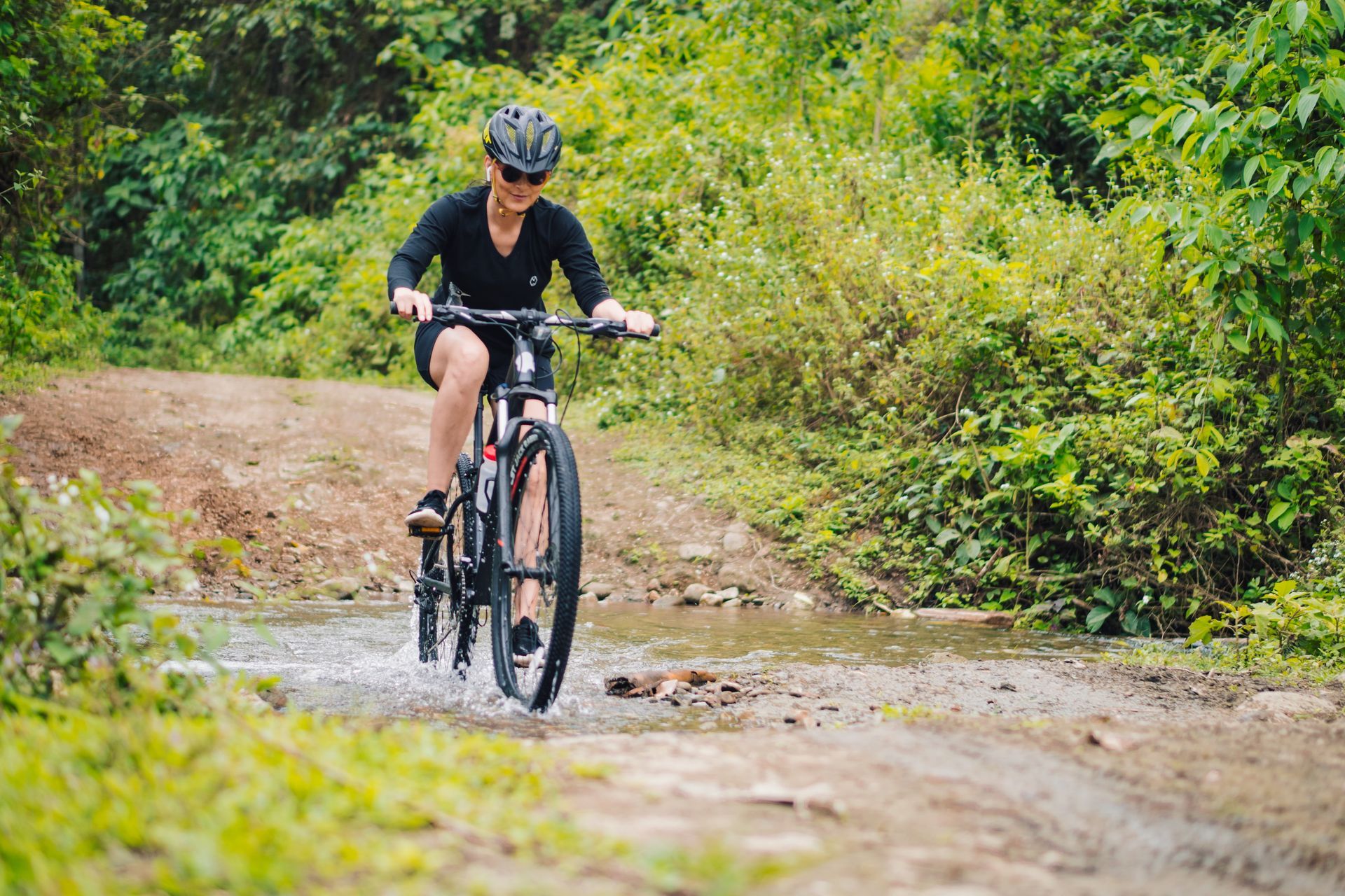 Woman on a mountain bike rides through a shallow stream on a dirt path in a forest.