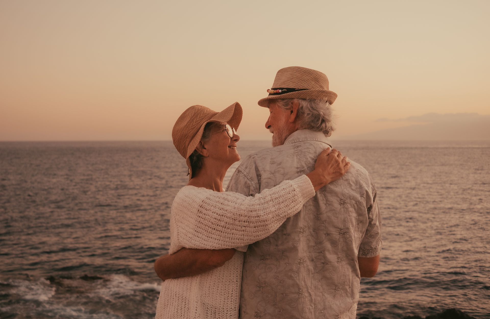 Elderly couple embracing, gazing at each other, ocean sunset.