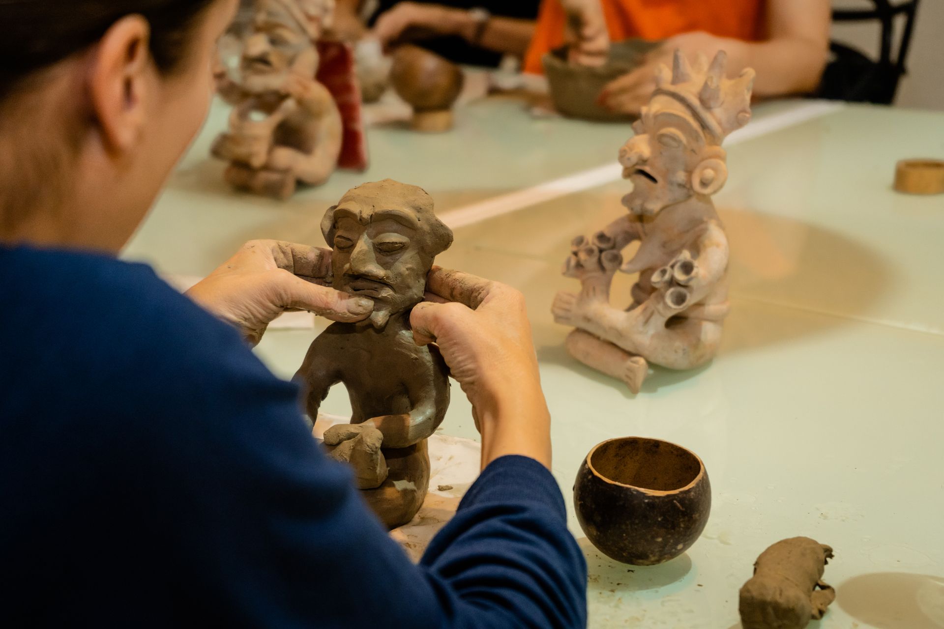 Person sculpting a clay figure, focusing on the face. Others work on similar figures at a table.