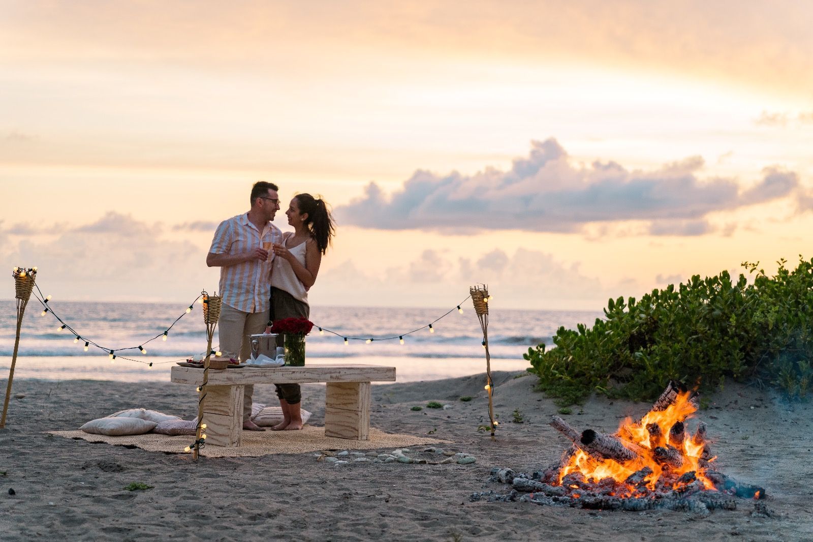 Couple toasting drinks on beach at sunset, lit by fire and string lights.