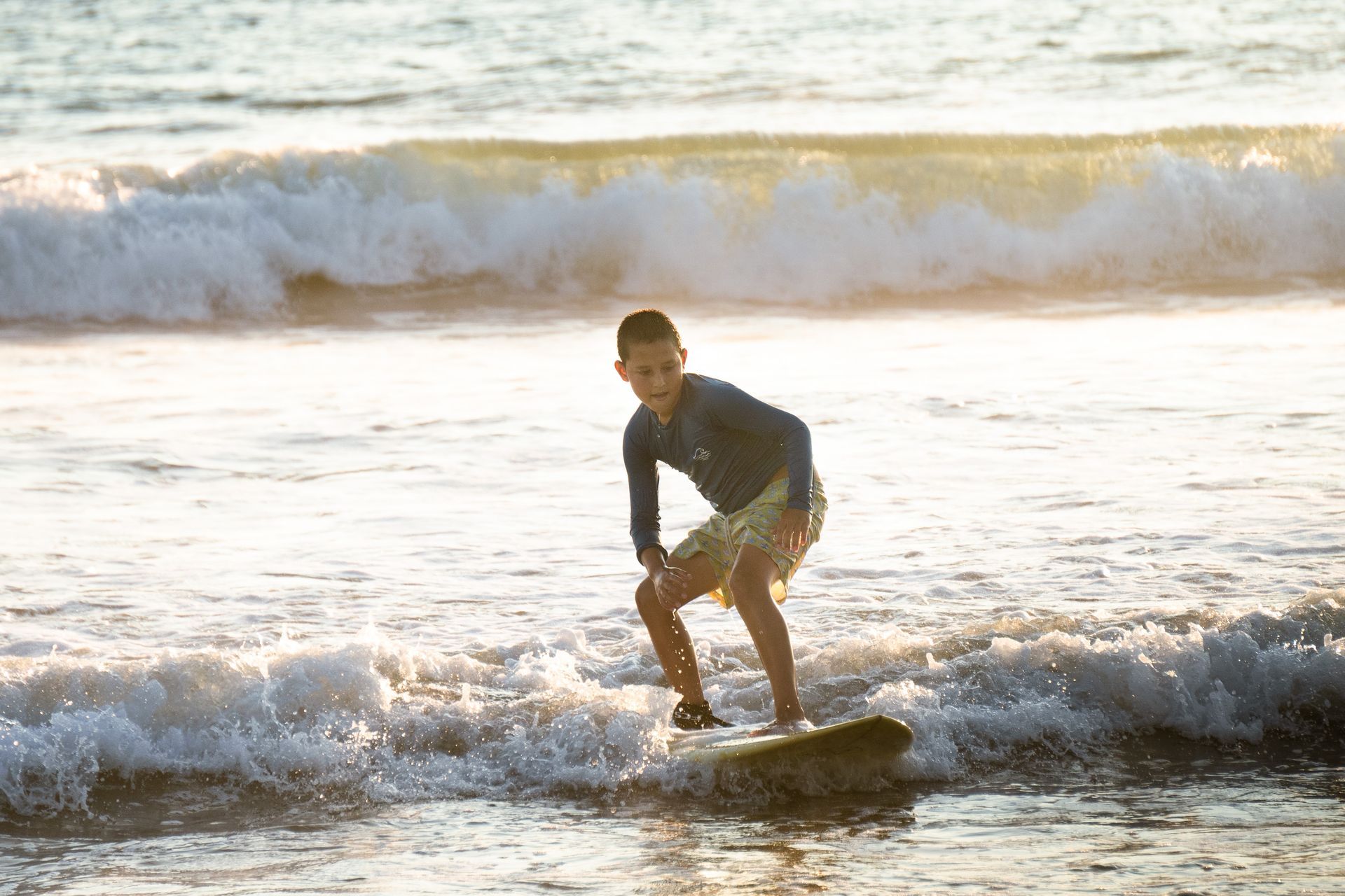 Boy surfing in the ocean, balanced on a board, smiling. Bright sun and waves in the background.