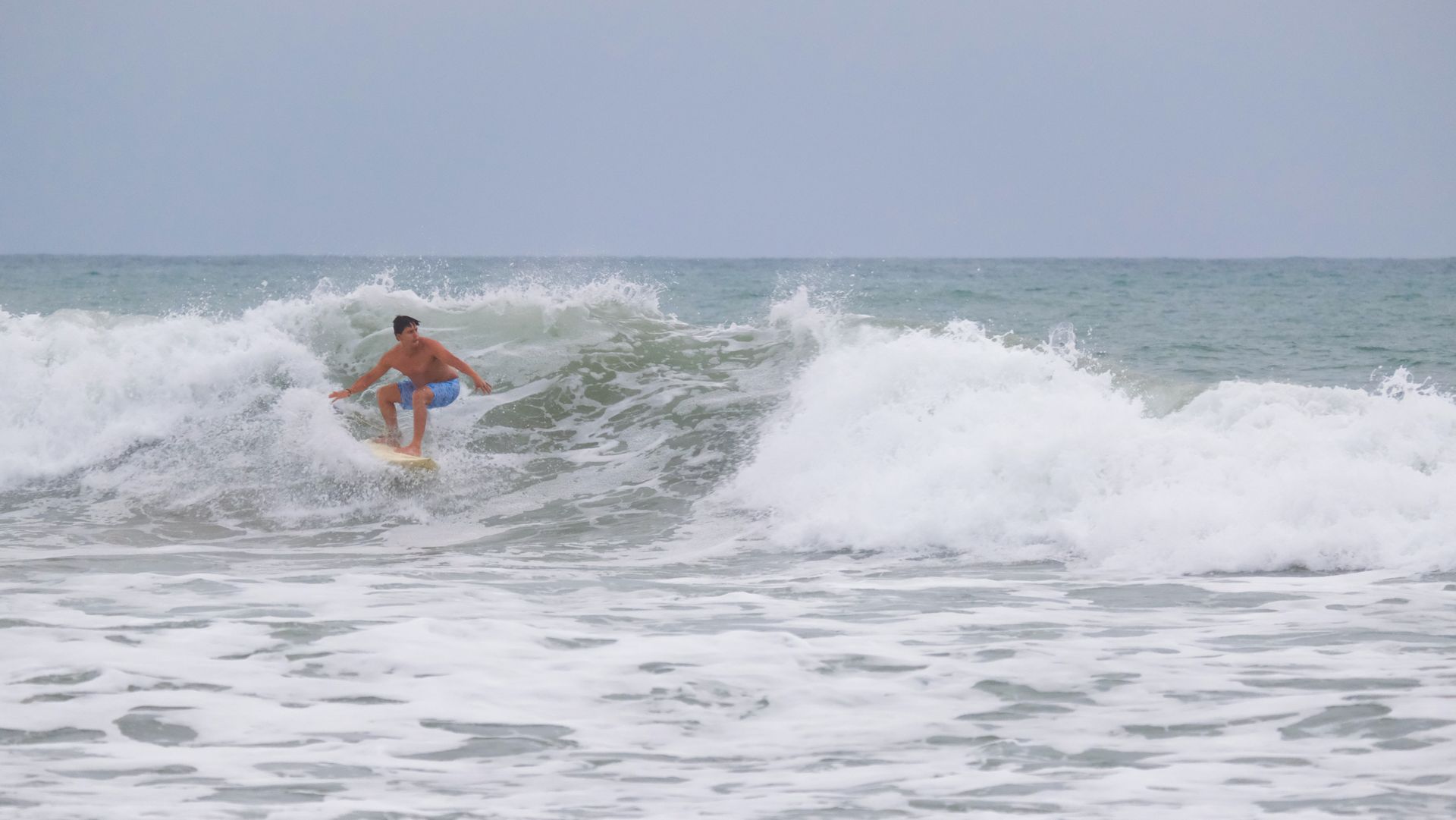 Surfer riding a wave in the ocean. Wearing blue shorts, overcast sky.