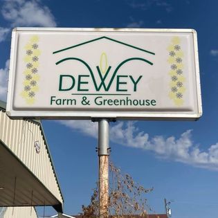 Sign for Dewey Farm & Greenhouse, white with green logo and text on a pole under a blue sky.
