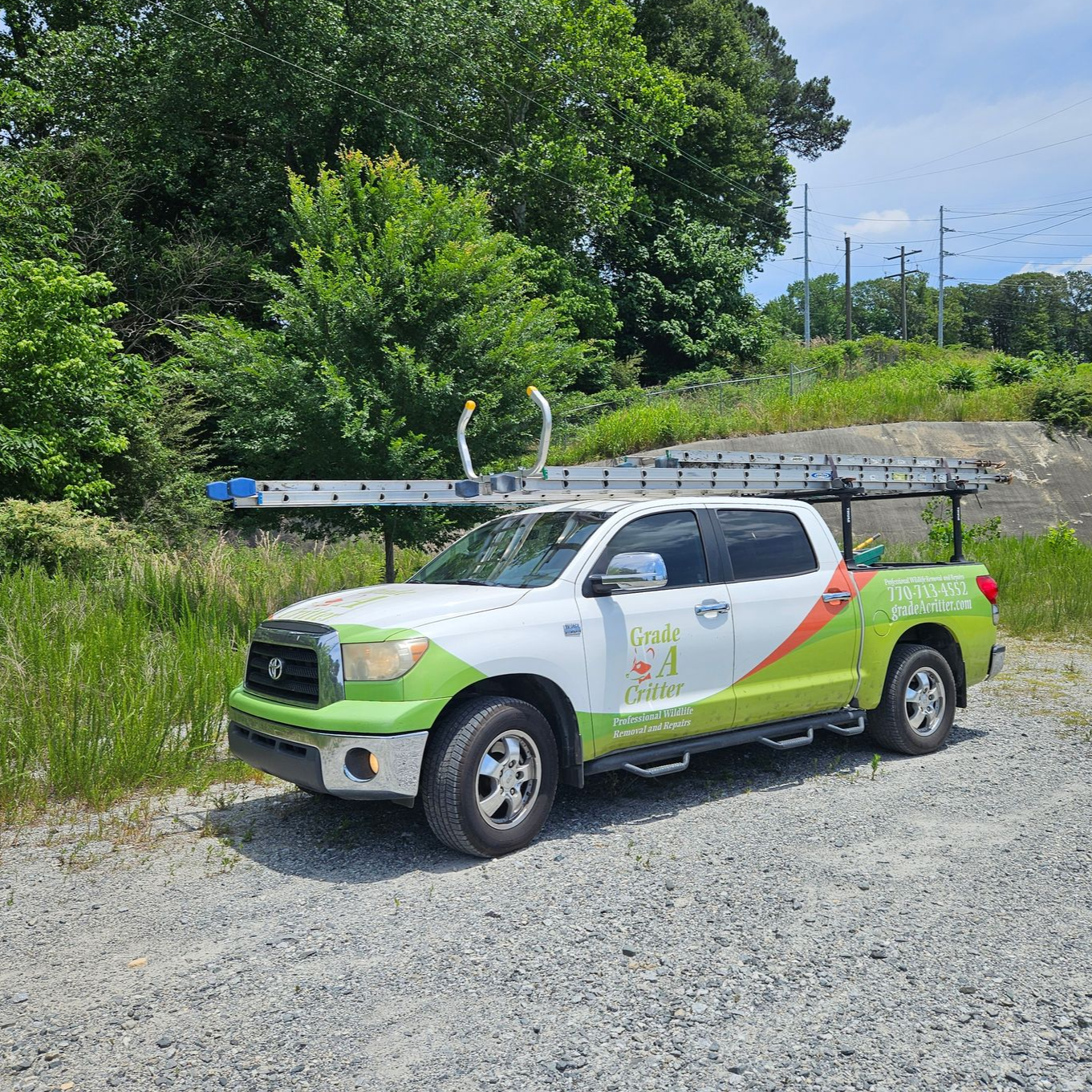 A white and lime green pickup truck with a ladder rack parked on a gravel lot near a wooded area.