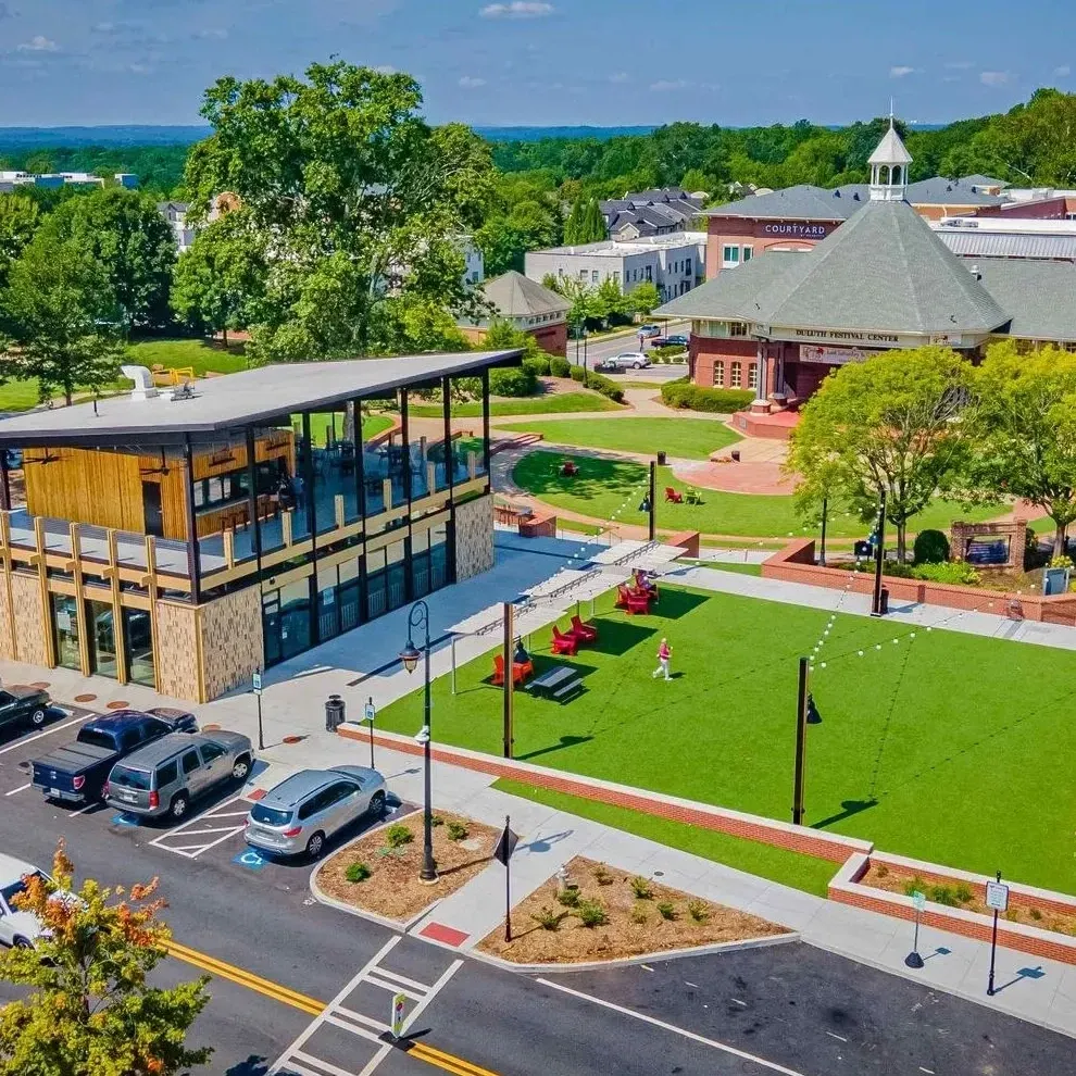 Aerial view of a campus with a modern building, green lawn, and a red brick pavilion near a road and parked cars