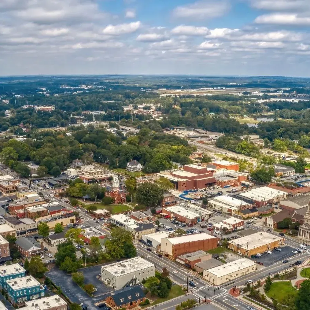Aerial view of a small town with trees, roads, low-rise buildings, and distant hills under a cloudy sky