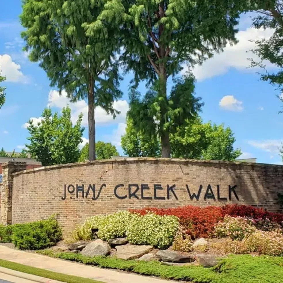Stone entrance sign reading “Johns Creek Walk” with landscaping and trees under a blue sky