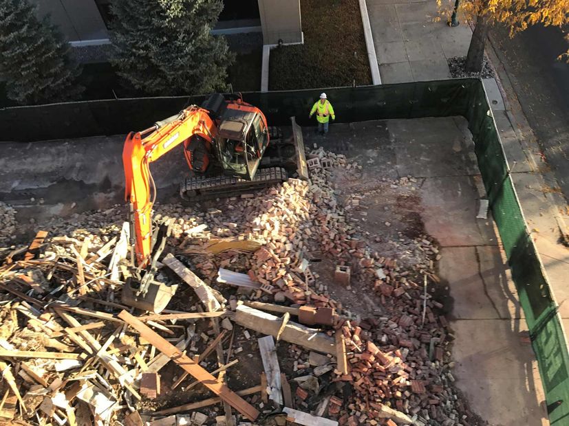 Excavator clearing debris in a walled lot, with a worker in a high-visibility jacket nearby.
