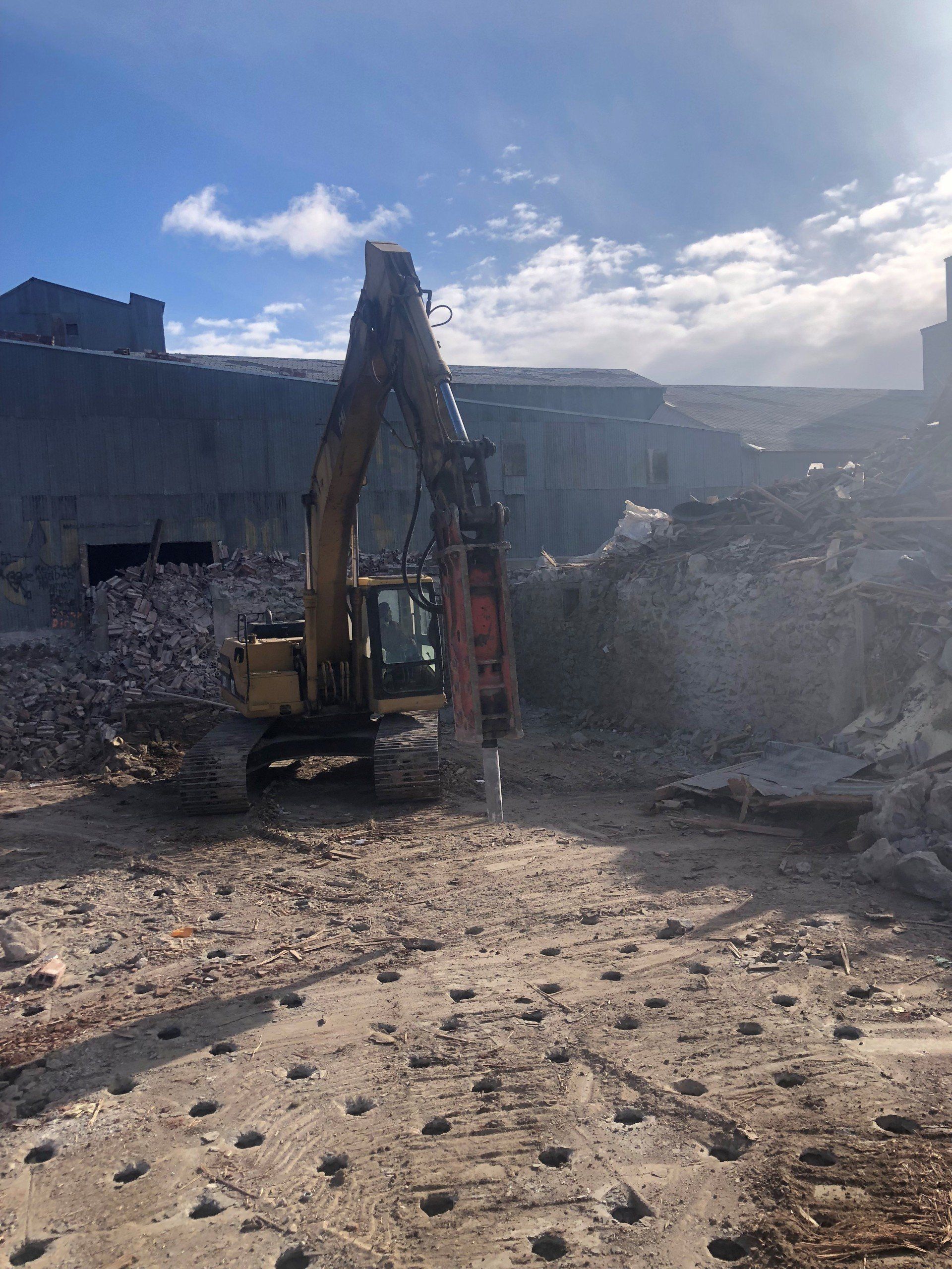 Excavator in a dusty demolition site, with rubble and concrete debris under a bright sky.