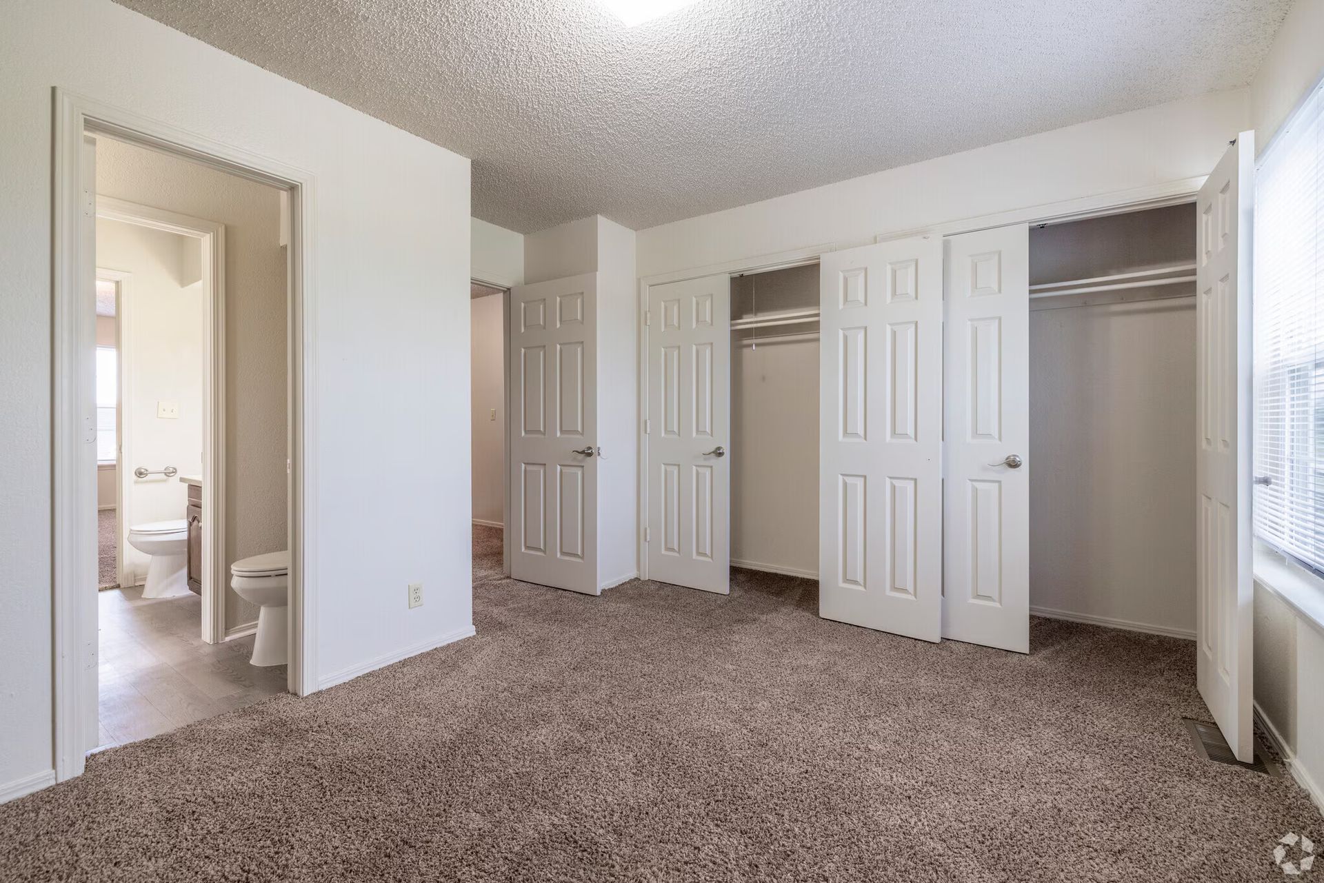 A bedroom with beige carpet, a textured ceiling, white walls, multiple closet doors, and an open doorway to a bathroom.