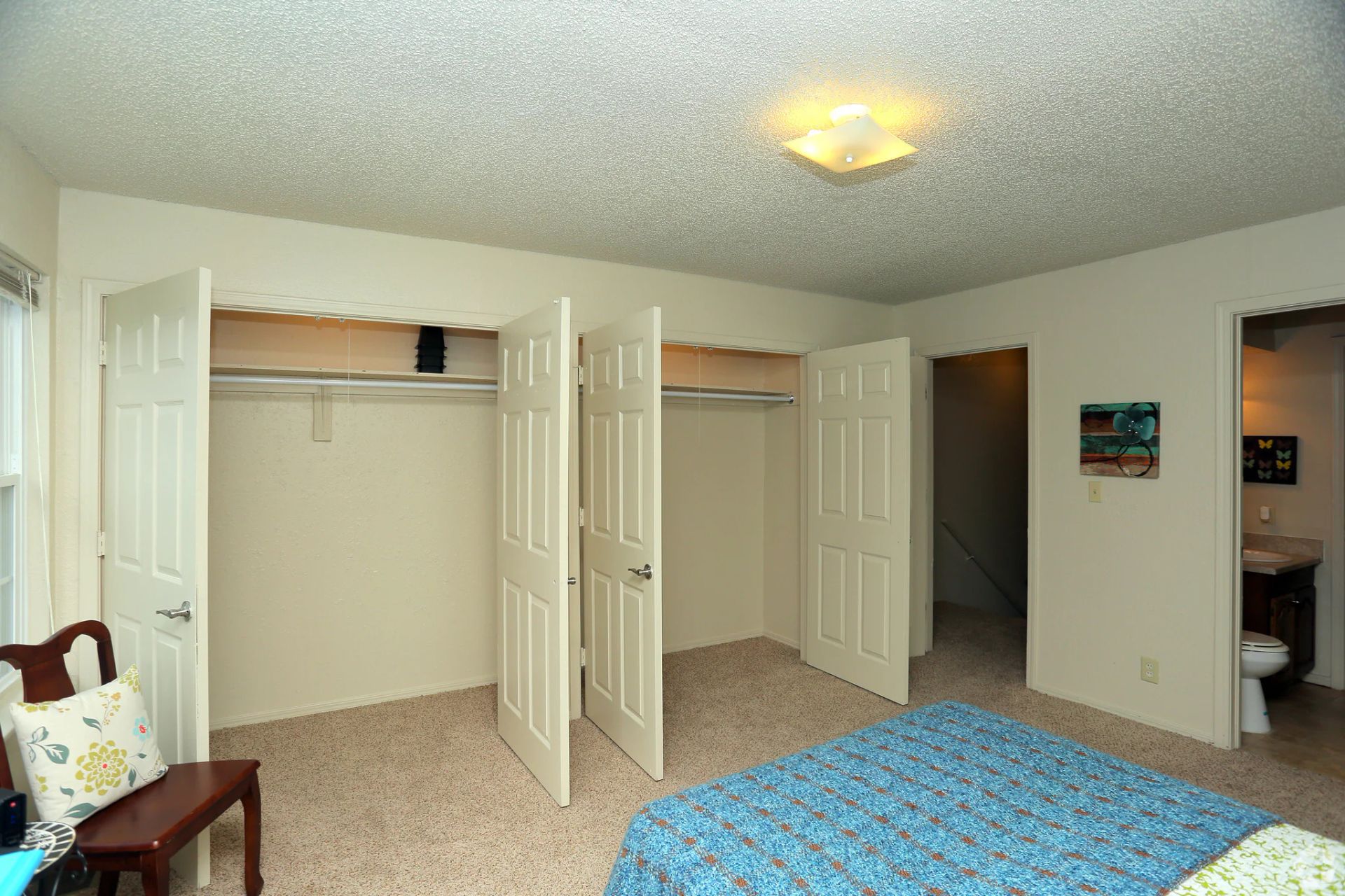 A bedroom interior featuring a blue patterned bed, wall-to-wall closets with white doors, and an attached bathroom entrance.