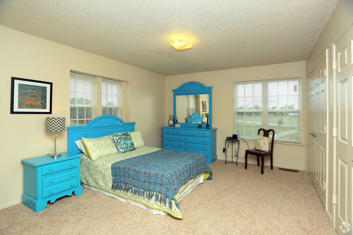 A bedroom with beige walls, a blue bed frame and furniture, a patterned throw blanket, and two windows with blinds.