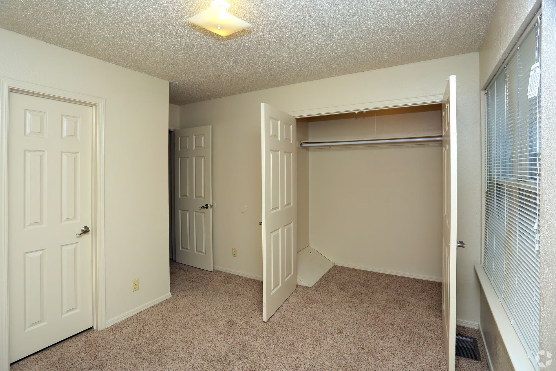An interior view of a bedroom with beige walls, tan carpeting, two white closed doors, and an open closet.