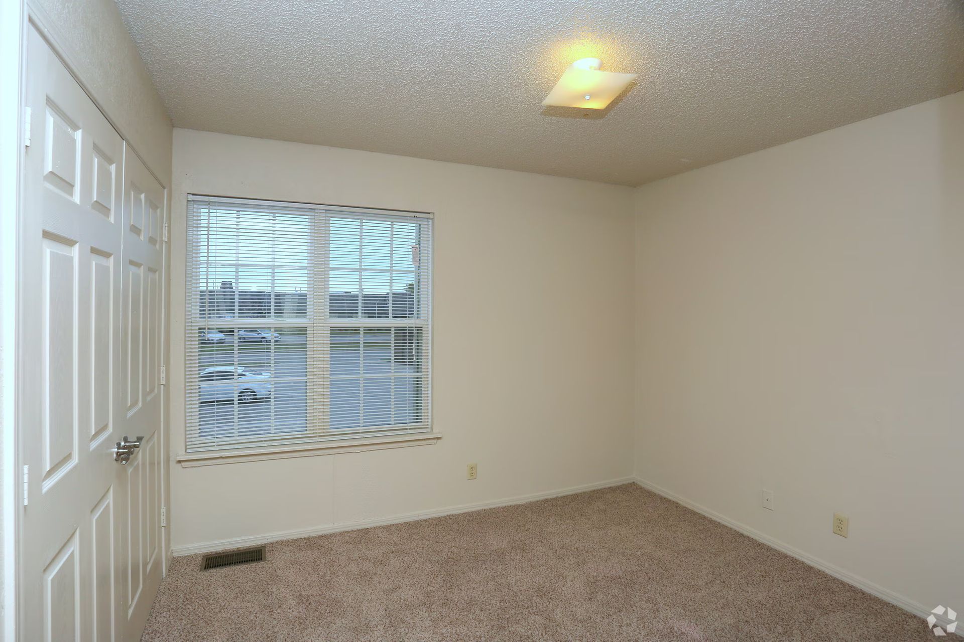 A small, empty room with beige carpeting, white walls, a closed white door, and a single window with horizontal blinds.