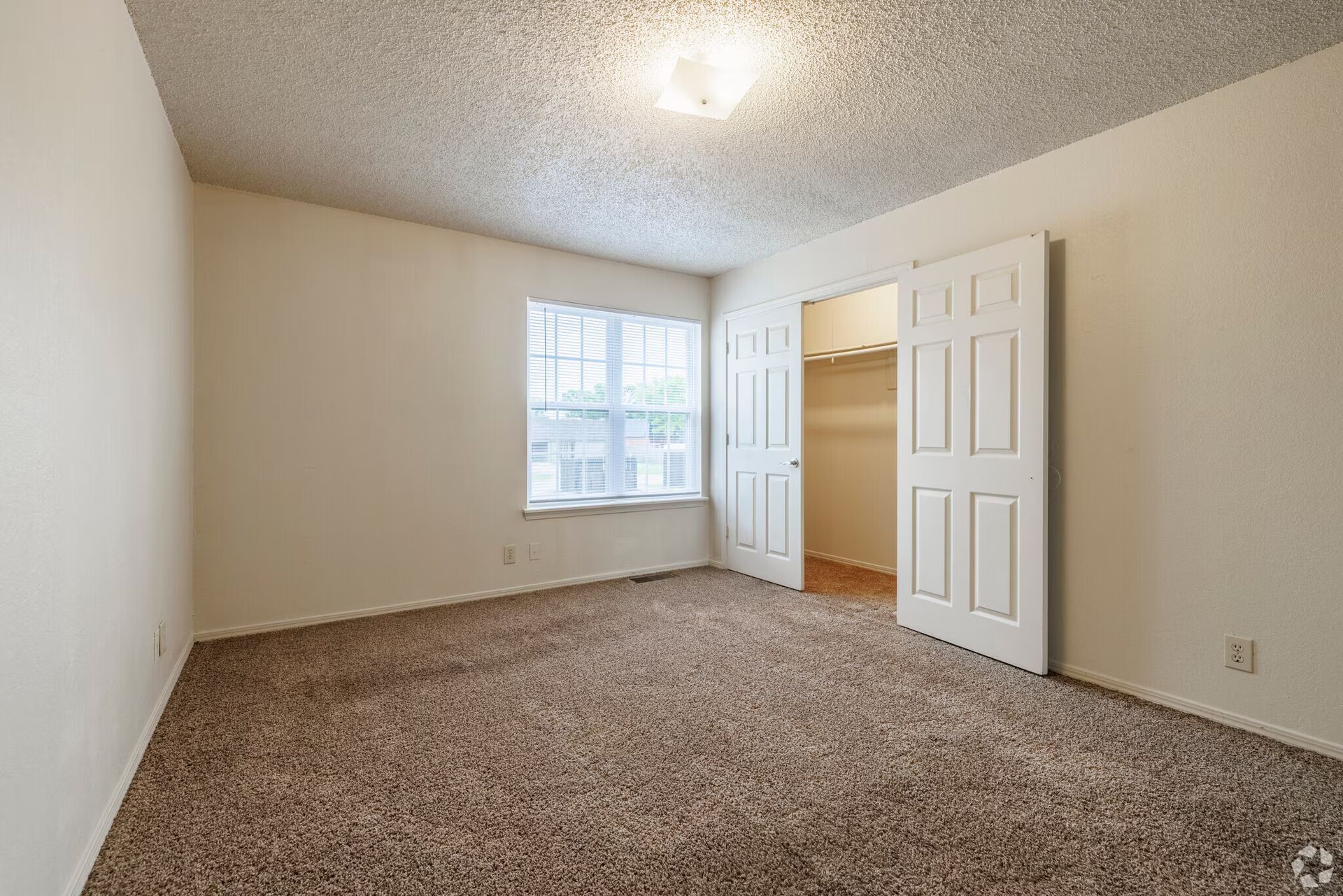 An empty bedroom with beige walls, brown carpet, a window, and an open door revealing a closet with shelving.