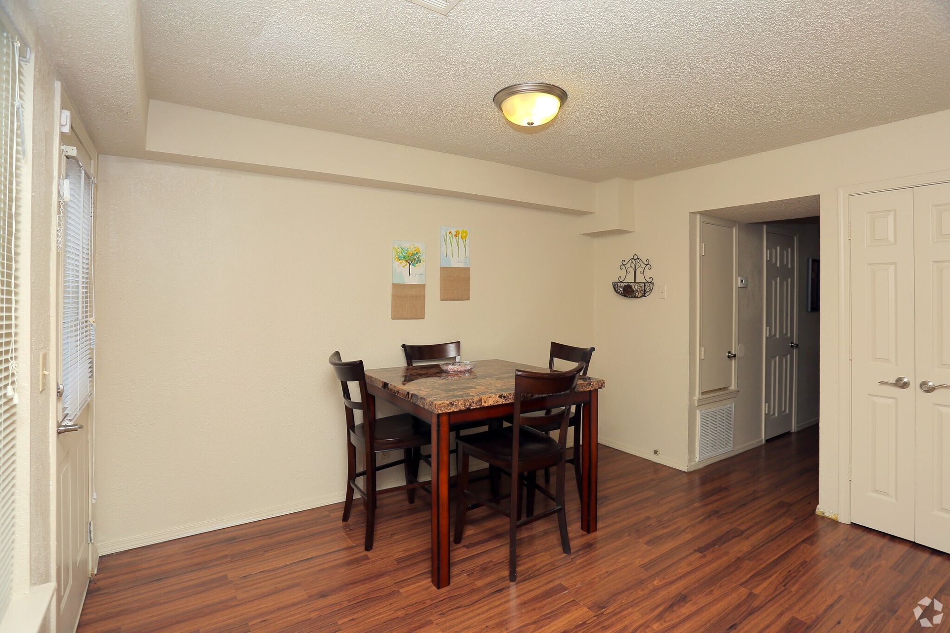A dining area with a dark wood table, four chairs, light walls, and wood-style flooring under a ceiling light.