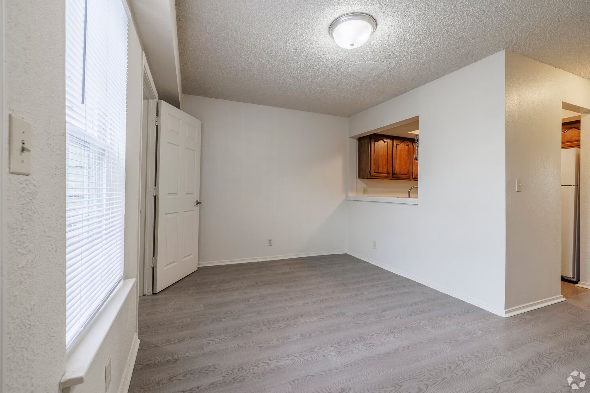A sunlit, unfurnished room with gray wood-style flooring, white walls, and a pass-through window into a kitchen.