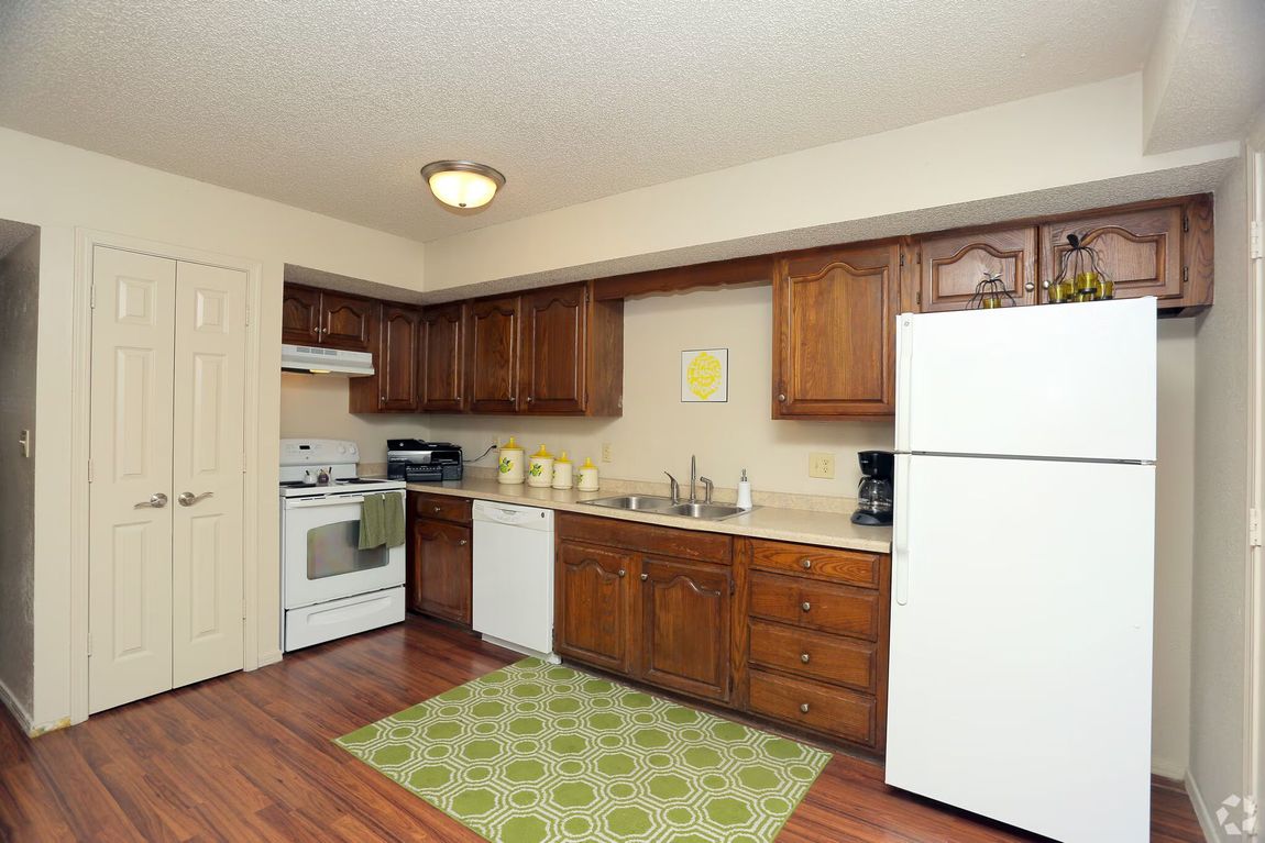 Kitchen with wood cabinets, white appliances, a light-colored countertop, and a green patterned rug on wood-style floor.