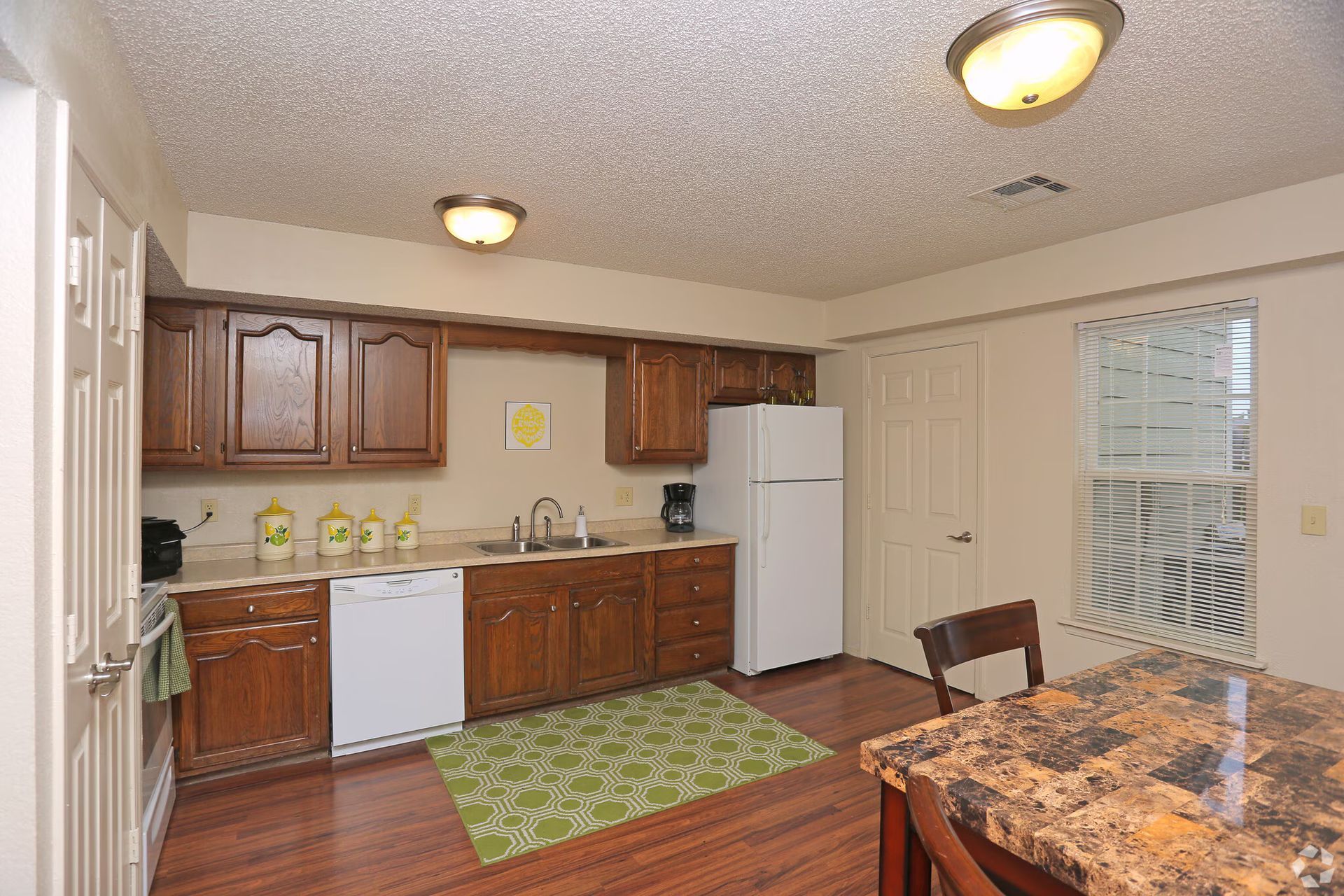A kitchen with dark wood cabinets, white appliances, brown countertops, and a green patterned rug on wood-look flooring.