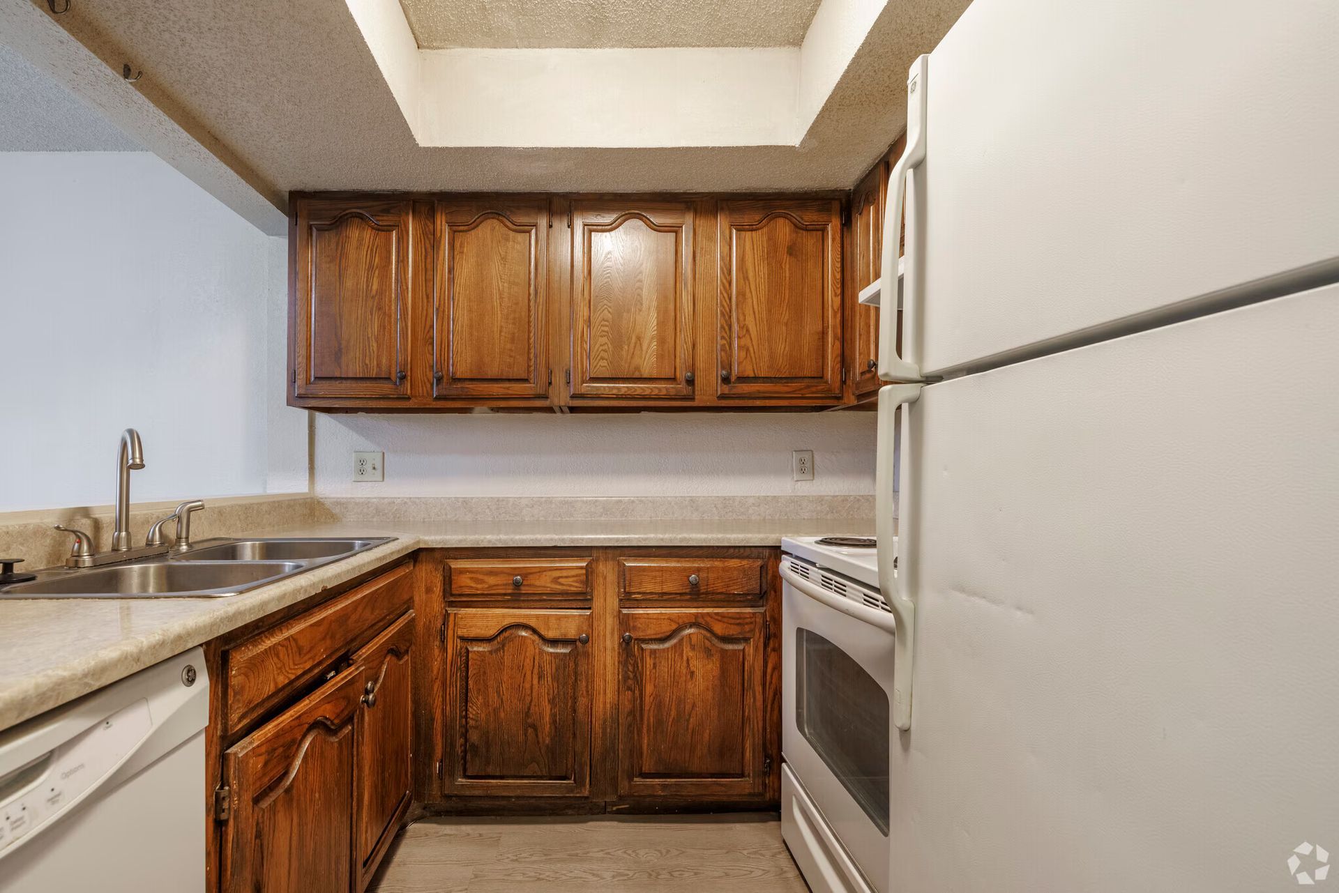 A kitchen with medium-toned wooden cabinets, beige countertops, a white refrigerator, and a stainless steel sink.
