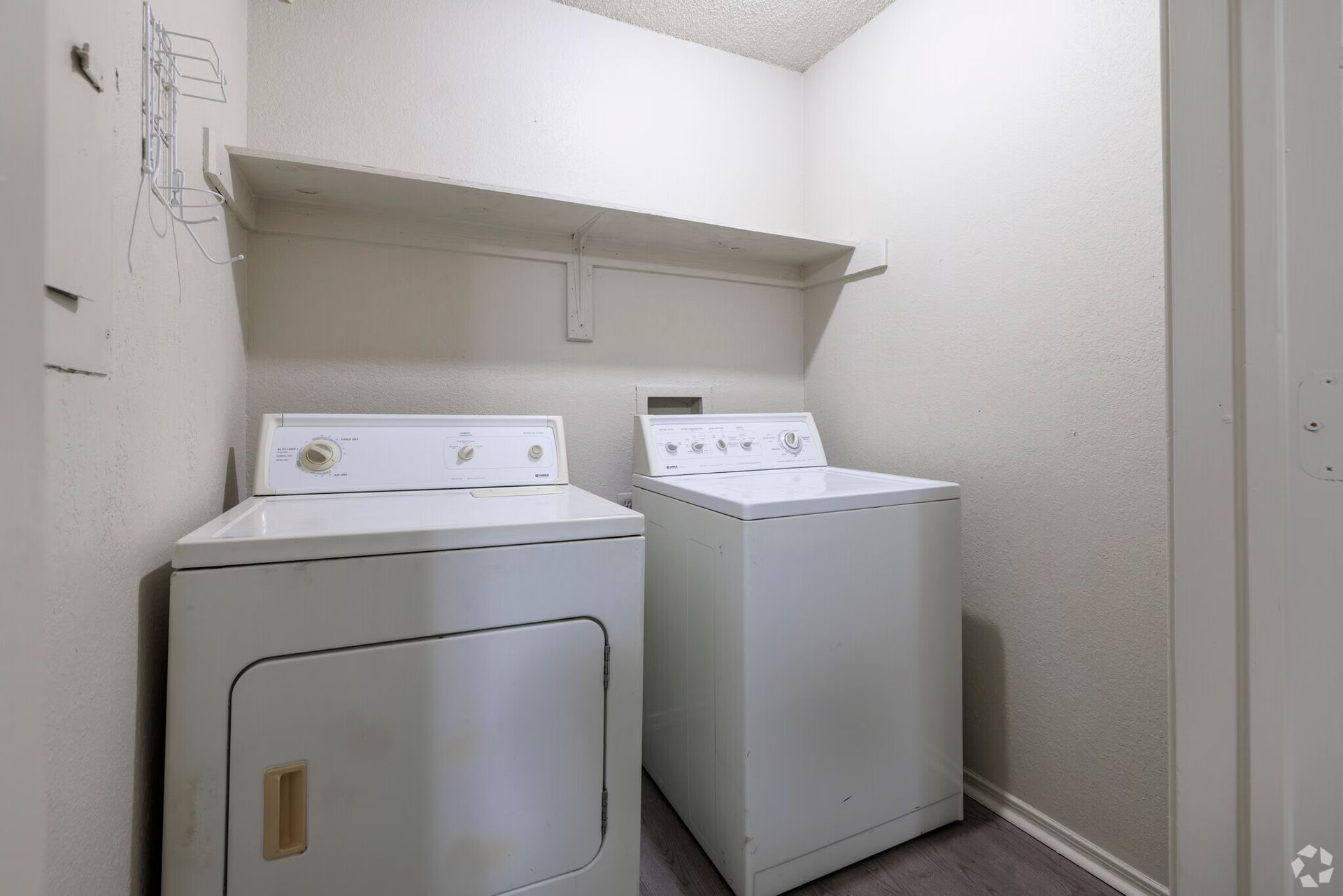 A laundry room with a side-by-side white washer and dryer under a wooden shelf against plain, off-white walls.