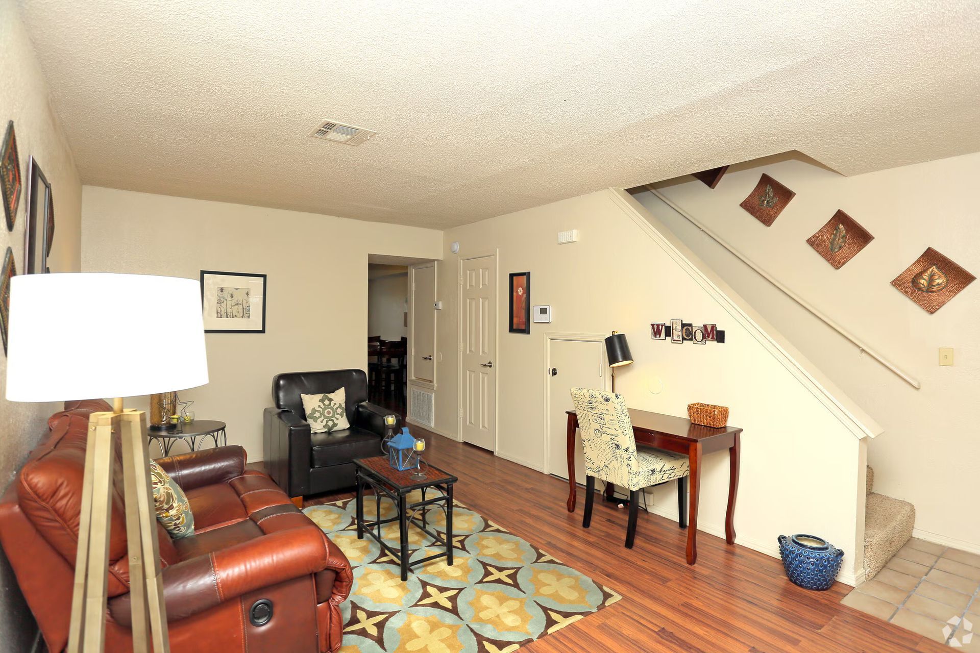A living area with a brown leather sofa, a black armchair, a small desk with a patterned chair, and stairs to the side.
