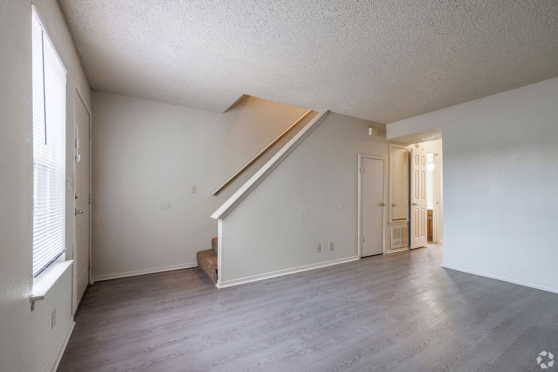 An empty living area with gray wood-style flooring, white walls, and a carpeted staircase leading to an upper level.