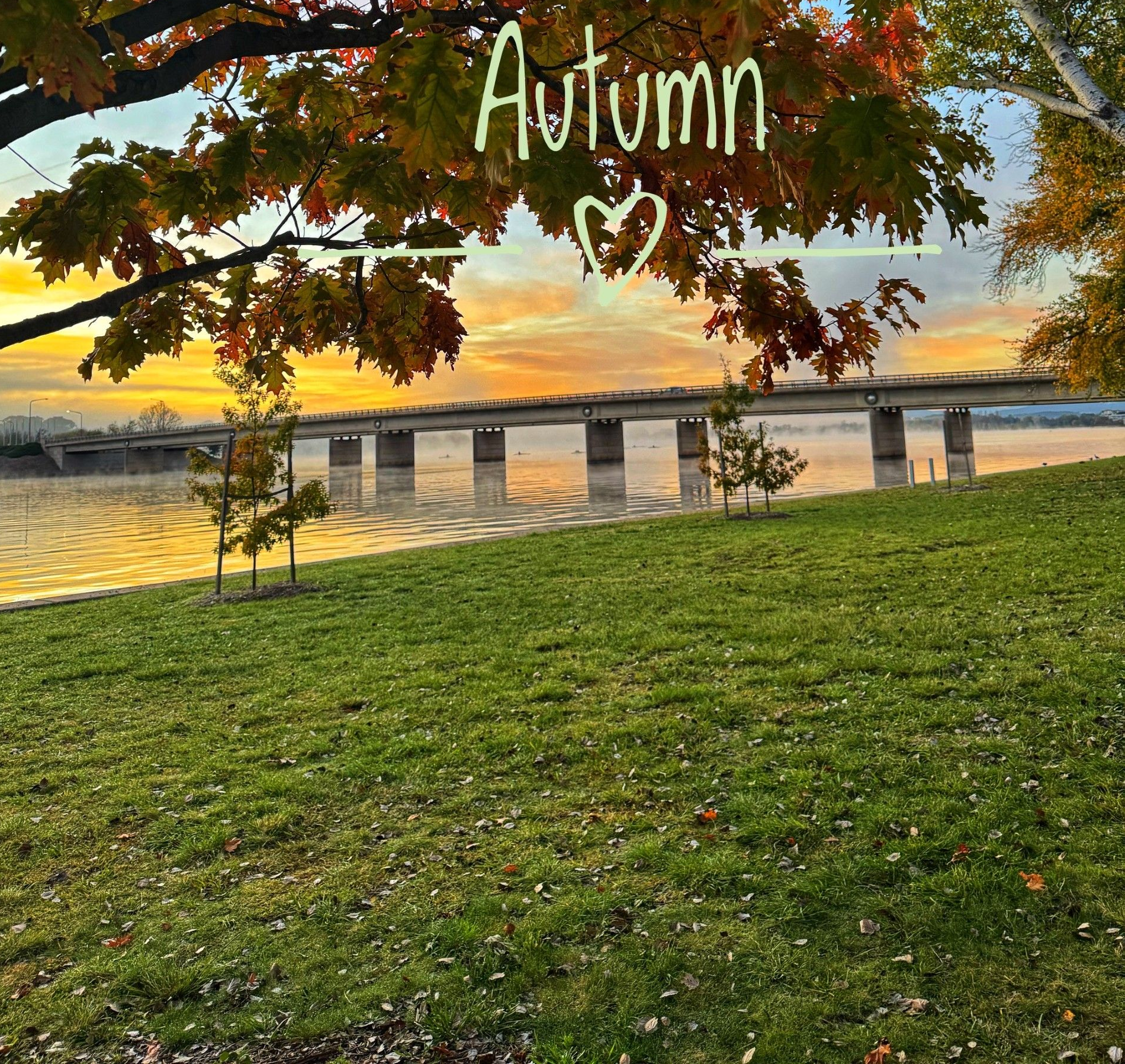 Autumn sunset over a river with a bridge, framed by orange leaves and green grass under a handwritten “Autumn” title.