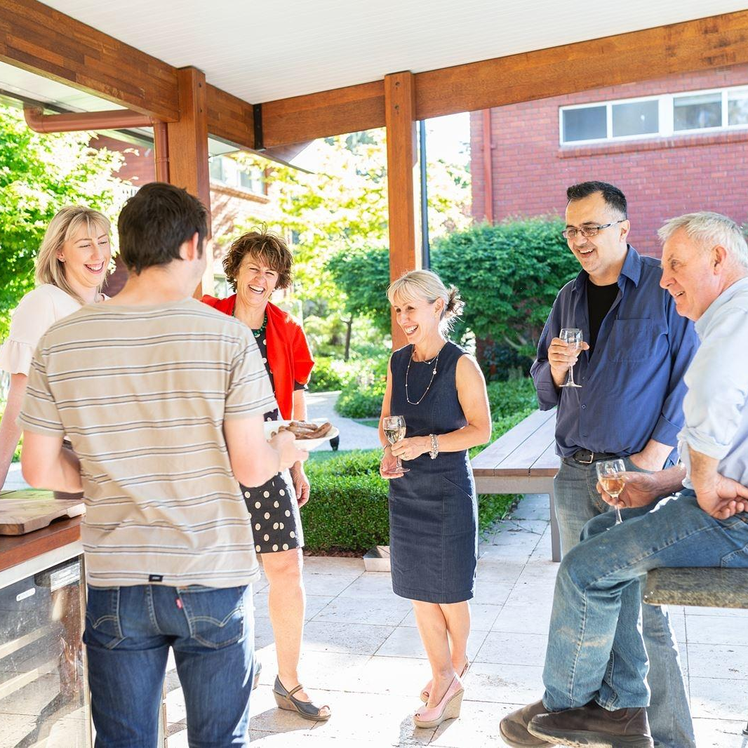 Group chatting and holding drinks under a wooden patio gazebo on a sunny day.