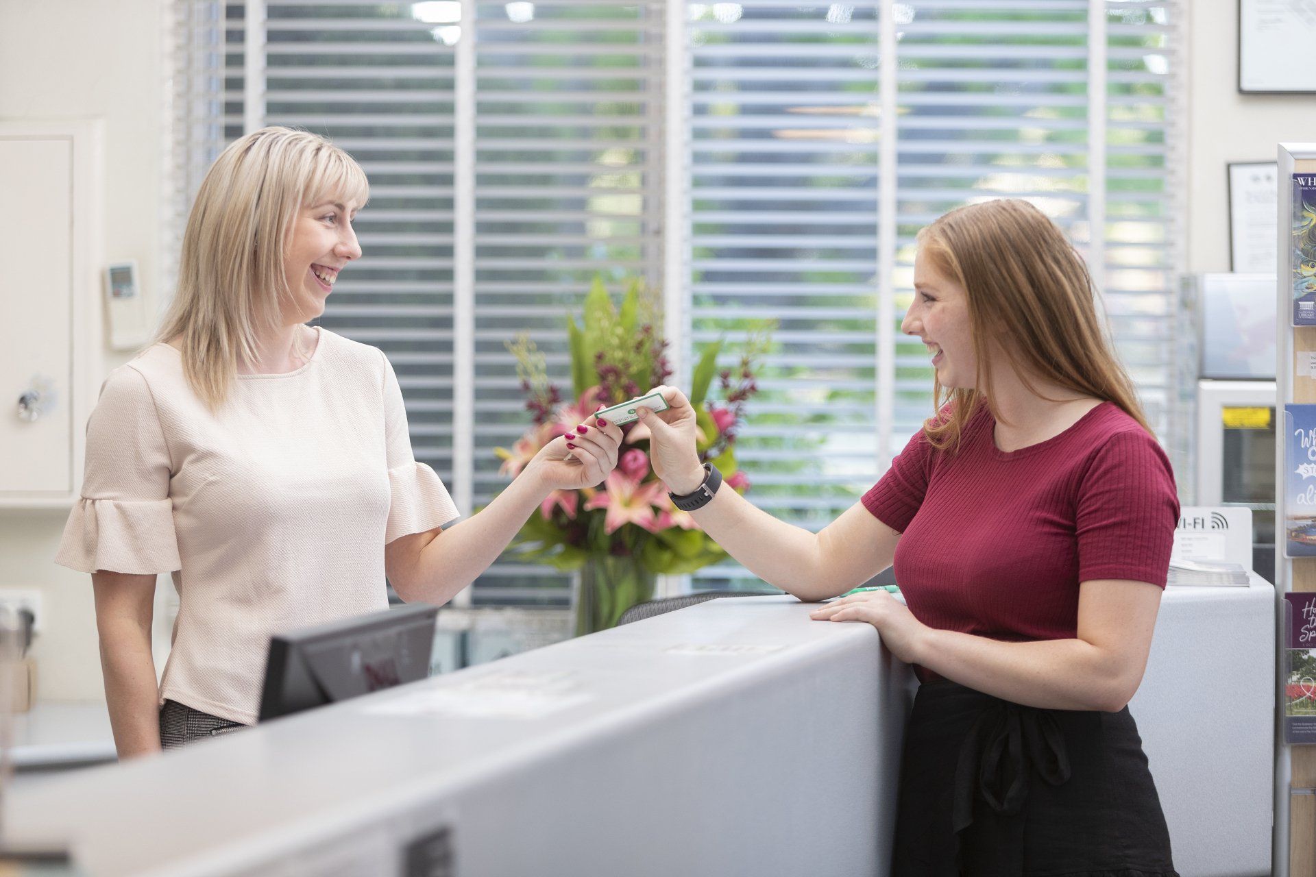 Two women exchange flowers at a reception desk in a bright office.